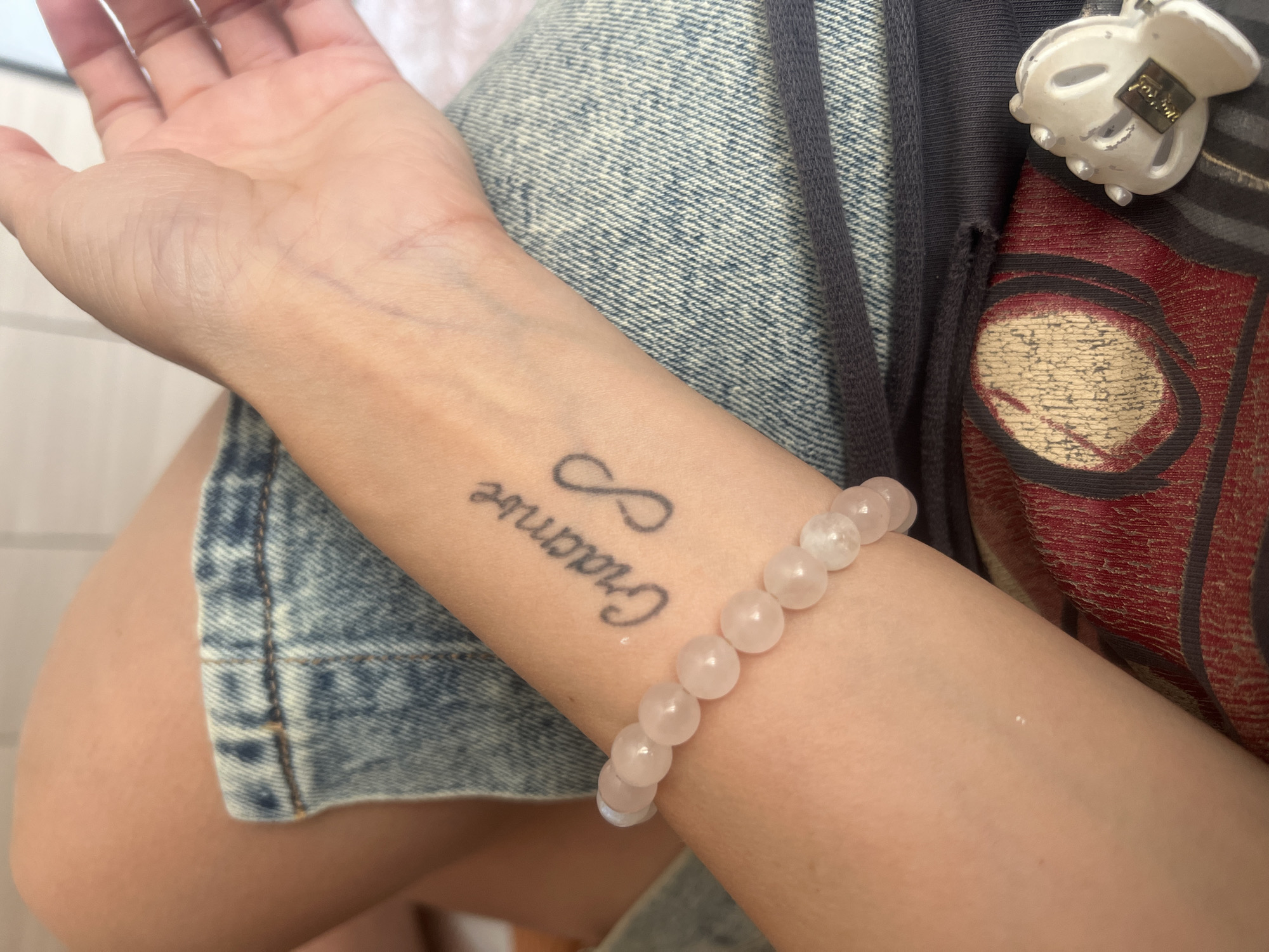 The image shows a close-up of a person's forearm and wrist, with a tattoo and a bracelet visible. The tattoo on the inner wrist reads "Счастье" in Cyrillic script, meaning "happiness," and is accompanied by an infinity symbol. A bracelet made of rose quartz beads is worn around the wrist. The person is wearing denim shorts and a dark-colored garment underneath. In the lower right corner, a white charm, possibly a keychain or part of a bag, is visible. The background suggests an indoor setting with soft lighting. The image is focused on the forearm, with other details blurred. The specific location within "Unknown, Unknown" cannot be determined from the visual information.