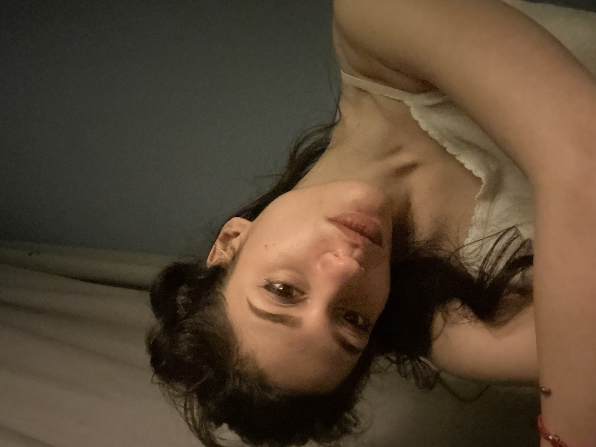 A young woman with dark, messy hair is shown in a close-up, head-and-shoulders shot. Her head is tilted to her right, and she is looking directly at the camera. She is wearing a white top with thin straps. The background is dark and out of focus, with a draped fabric on the left side. The lighting is dim and appears to be coming from the right. The image is a selfie. The location is not specified in the image or its metadata.