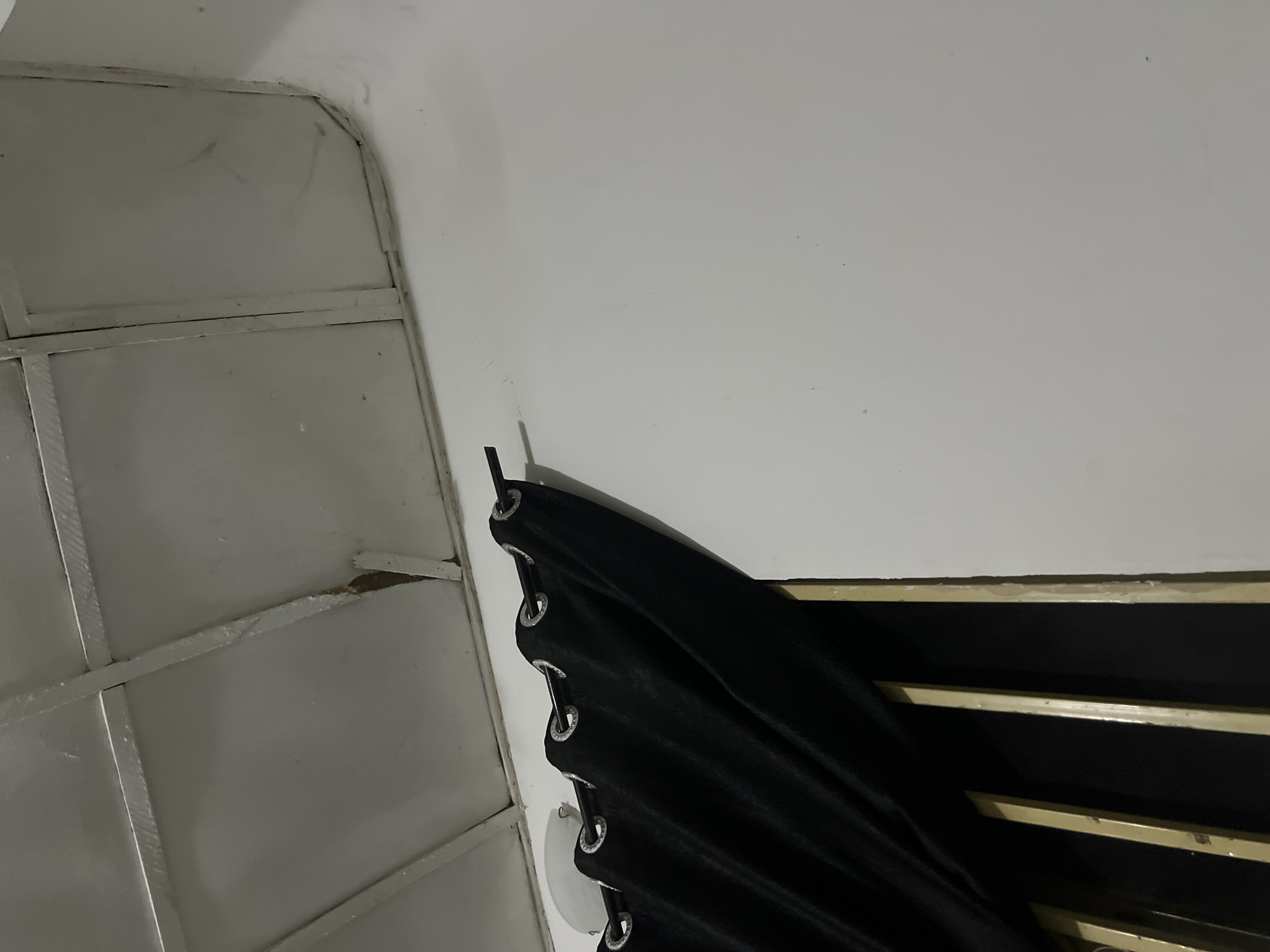 The image depicts an interior scene, likely a room. A black curtain with silver grommets hangs from a rod that is attached to the wall. The curtain is drawn to the left, revealing some vertical bars behind it, possibly part of a window or a grill. The ceiling appears to be made of panels and shows some signs of wear or damage, with a piece of the material hanging loose in one section. The walls are painted a light color, possibly white or off-white, and there are some faint marks or stains visible on the wall surface. The lighting suggests it is either daytime with artificial lighting or evening. There are no people visible in the image. The scene does not contain any specific indicators to suggest a location within the city of Howayi, Nigeria.