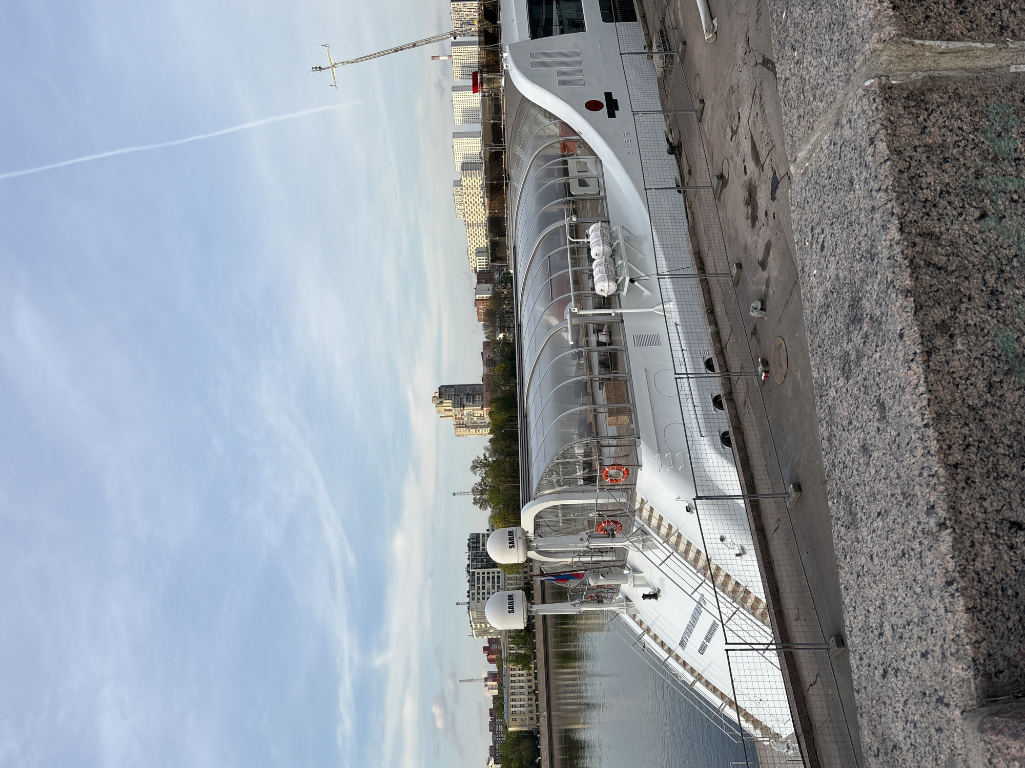 The image depicts a portion of a white river cruise ship docked on the embankment in Saint Petersburg, Russia. The ship is long and sleek, featuring a distinctive enclosed passenger area with clear windows. It has two large, white radomes labeled "SAILOR" on the top deck. A Russian flag is visible. The ship is docked along a concrete embankment, with a section of granite curb and sidewalk visible in the foreground. A temporary metal fence separates the ship from the pedestrian area. In the background, city buildings of varying heights and styles can be seen, including residential buildings, office buildings, and construction cranes. The sky is partly cloudy, with a faint contrail. The water in the canal is calm. No people are directly visible in the shot.