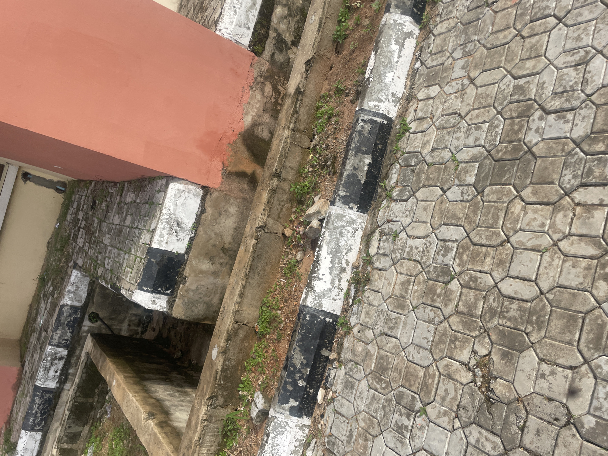 This is a forensic, matter-of-fact description of an outdoor scene in Lokoja, Nigeria.
The image depicts an exterior environment with concrete structures and paving. In the foreground, there is a textured grey interlocking paver surface, appearing wet and slightly worn in places. Adjacent to this is a raised concrete curb, painted with alternating black and white stripes that are faded and chipped, indicating age and exposure to the elements. This curb borders a shallow, open concrete channel or drain. Beyond this channel, another raised concrete structure is visible, also topped with the same black and white striped border. This structure is adjacent to a larger, rust-colored concrete pillar or wall, which takes up a significant portion of the right side of the frame. The pillar shows signs of water staining and some discoloration near its base.
To the left of the main concrete channel, there appears to be a deeper, open concrete pit or trench, with visible debris and some sparse green vegetation growing within it and around its edges. The ground surrounding the concrete structures is composed of soil and small stones, with patches of weeds and grass growing in scattered areas. The overall impression is one of an outdoor utility or drainage area, possibly part of a public space or building's exterior, showing signs of weathering and disrepair. No people or animals are present in the image. The lighting suggests an overcast or shaded day.