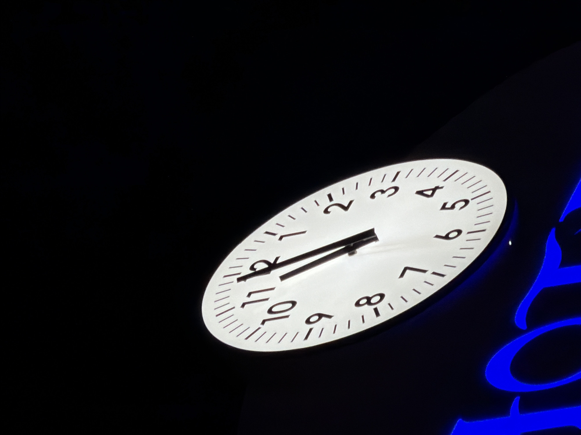 The image shows a close-up, low-angle shot of a large, illuminated clock face against a dark background. The clock has Roman numerals and minute markers, with the hour hand pointing slightly before the 11 and the minute hand pointing at the 12. To the left of the clock, partially visible at the bottom, are bright blue, illuminated letters that spell out "HOTEL". The overall scene suggests a hotel sign at night. The location is identified as Gatteo, Italy.
