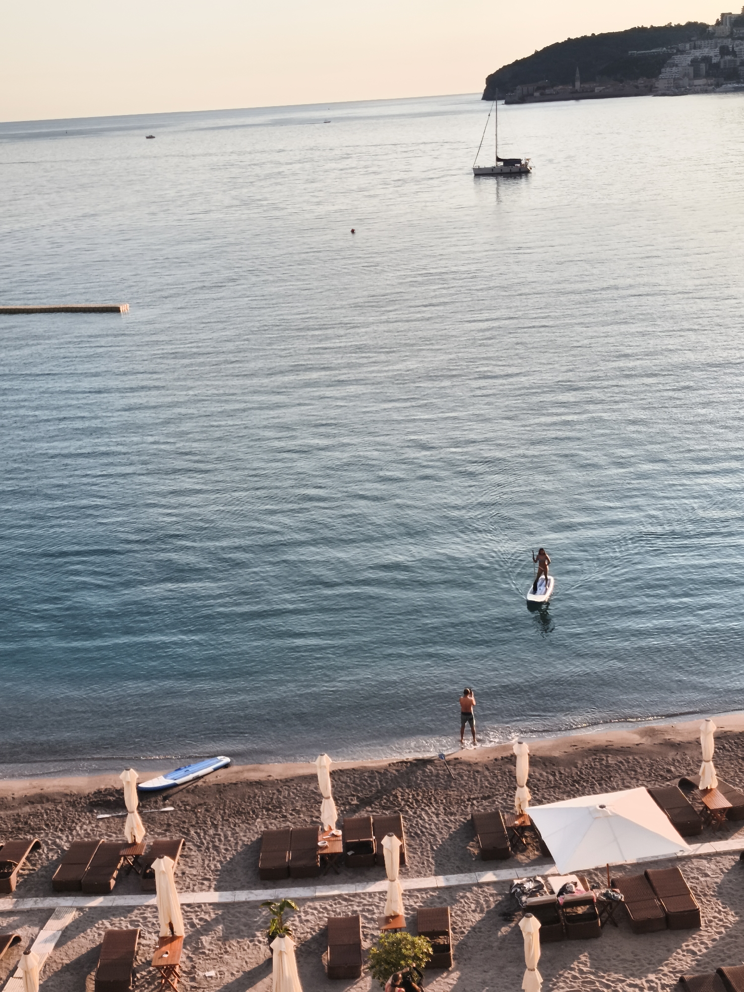 A high-angle shot captures a tranquil coastal scene in Budva, Montenegro, likely during the golden hour or late afternoon. The image is divided between the calm, blue-green sea and a sandy beach.
In the mid-ground, a person on a paddleboard glides across the water, actively paddling. Further out, a sailboat with its mast visible is moored, and several smaller vessels are scattered across the vast expanse of the sea. A short, wooden pier or dock segment extends from the left into the water. The horizon features a distant, hilly coastline densely covered with buildings, characteristic of the Montenegrin coast.
The foreground showcases a sandy beach with an array of amenities. Numerous dark-framed sun loungers are arranged in rows, accompanied by tall, light-colored parasols casting long shadows due to the low sun angle. On the sand near the water's edge, a blue and white inflatable paddleboard lies next to a paddle. Another person stands at the very edge of the water on the beach, looking out towards the sea.
The lighting is soft and warm, contributing to a peaceful atmosphere. The sky is clear, transitioning from a pale blue overhead to a lighter, almost peach hue near the horizon. No discernible text is visible within the image.