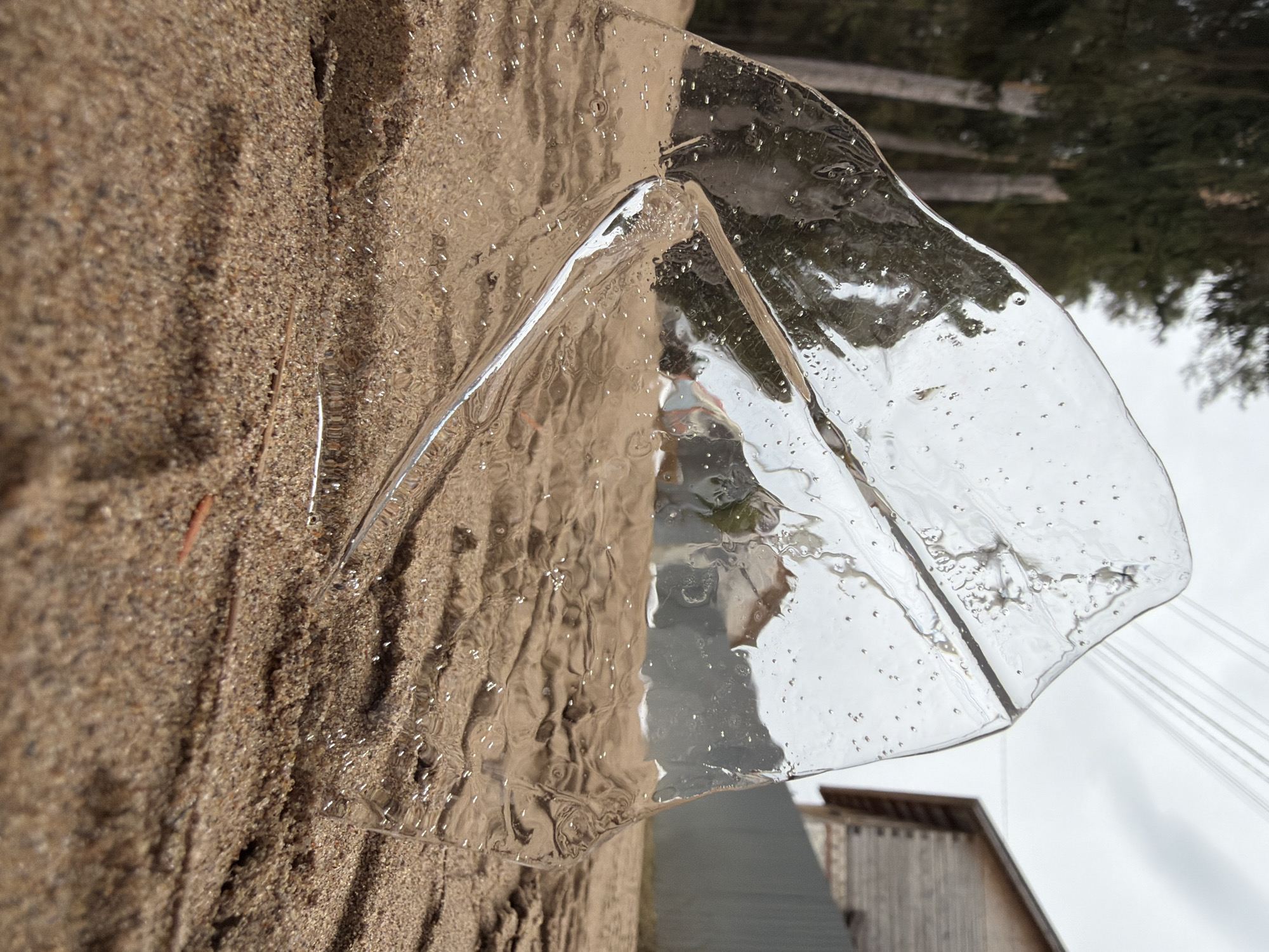 A warped sheet of clear ice lays on a sandy surface, partially obscuring a view of trees, a building, and overhead cables. The ice reflects the colors of the sand and the sky, and the overall scene appears to be outdoors, in daylight. There is no evidence to confirm the location is in the city of Chaikovsky, Russia.
