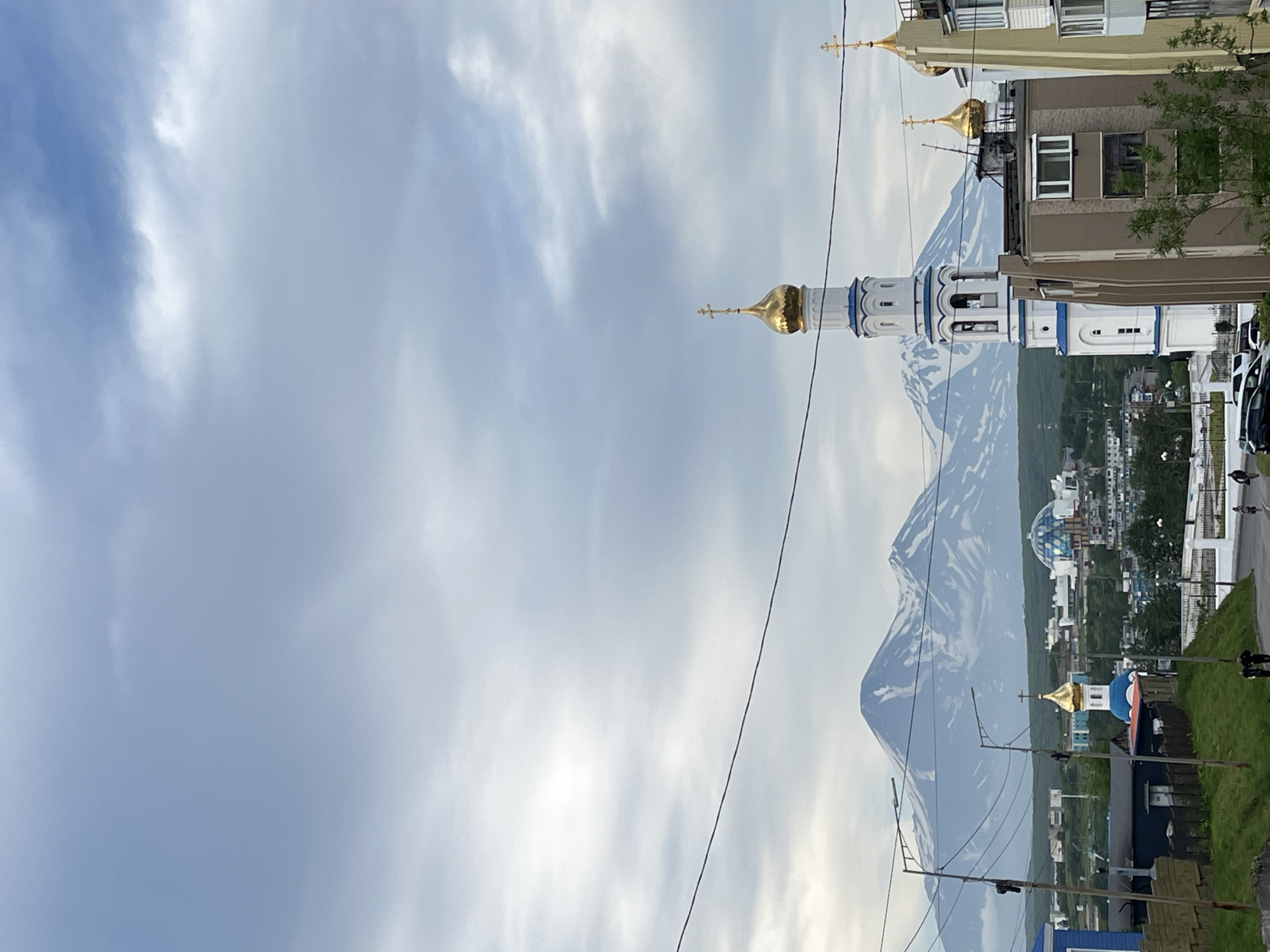 The image depicts a view of Petropavlovsk-Kamchatsky, Russia, with prominent volcanoes in the background. In the foreground, a white bell tower with a golden dome and a cross stands to the right of a modern building. Further to the left, a smaller blue-domed church with a golden cross is visible, nestled amongst other buildings and greenery. The sky is overcast with diffused light. Several power lines run horizontally across the upper portion of the frame. In the mid-ground, a cluster of buildings forms part of the city's urban landscape, with residential and commercial structures. The distant volcanoes, partially snow-capped, loom majestically behind the city. In the lower portion of the image, a paved road is visible, with a few people walking and some parked vehicles. The overall scene captures a blend of urban life and the dramatic natural environment characteristic of the Kamchatka region.