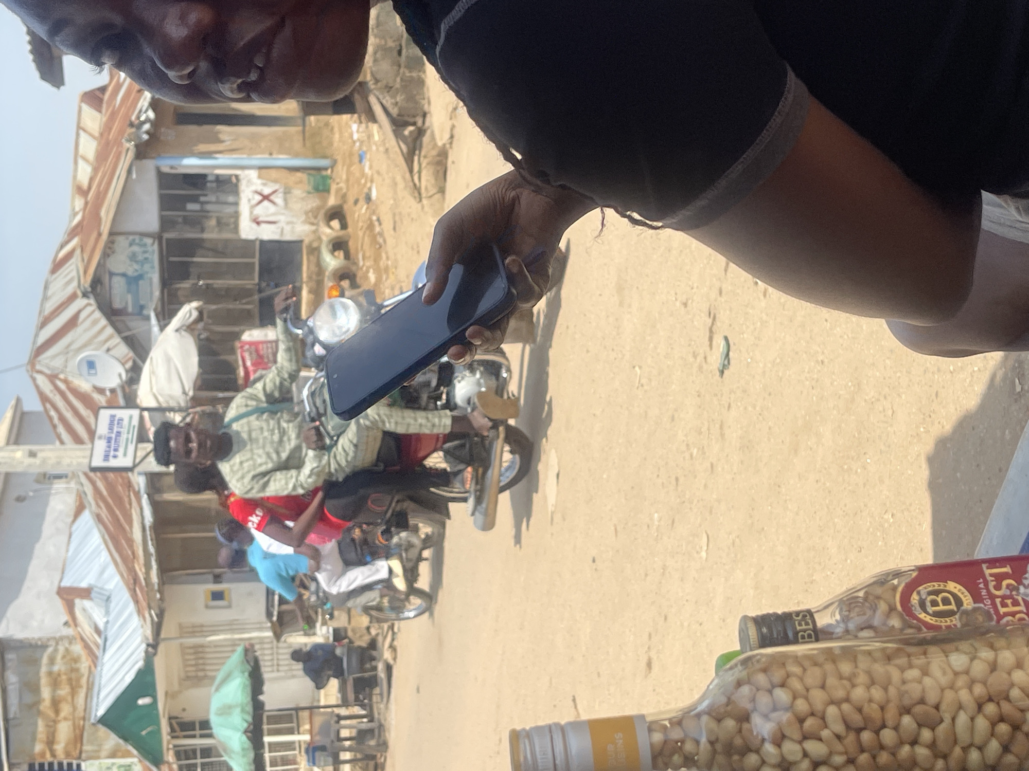 In a street scene in Abuja, Nigeria, a person in the foreground is holding a smartphone. Behind them, a man wearing a patterned long-sleeved shirt rides a motorcycle. Another person wearing a red shirt is seated behind him, and a third person in a blue shirt is riding on the back. The motorcycle is moving on a dirt road in front of a row of buildings with corrugated metal roofs. In the lower left of the image, two bottles are visible, one containing what appears to be nuts and the other a reddish liquid with a label that reads "BEST." The environment suggests a bustling marketplace or a residential area with commercial activity.