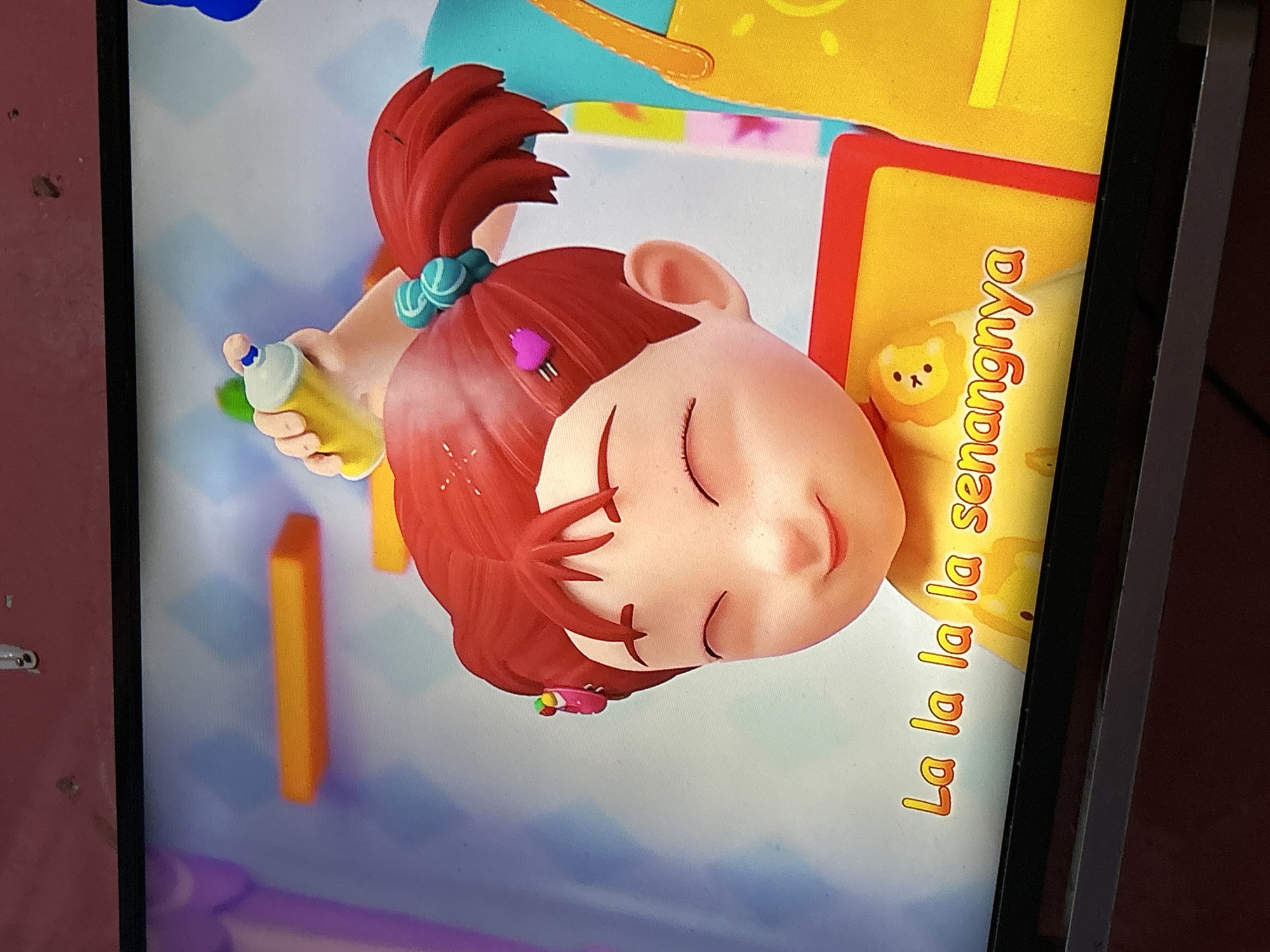 The image shows a close-up of a cartoon girl with red hair, wearing a yellow shirt with a lion graphic. She has her eyes closed and a smiling expression. The text "La la la la senangnya" is visible in orange lettering on the lower part of the image. In the background, a hand is holding a white spray can with a blue cap, and there is a hint of a blue and yellow apron. The setting appears to be a brightly colored, possibly indoor, environment. There is no identifiable information to suggest a location in Depok, Indonesia.