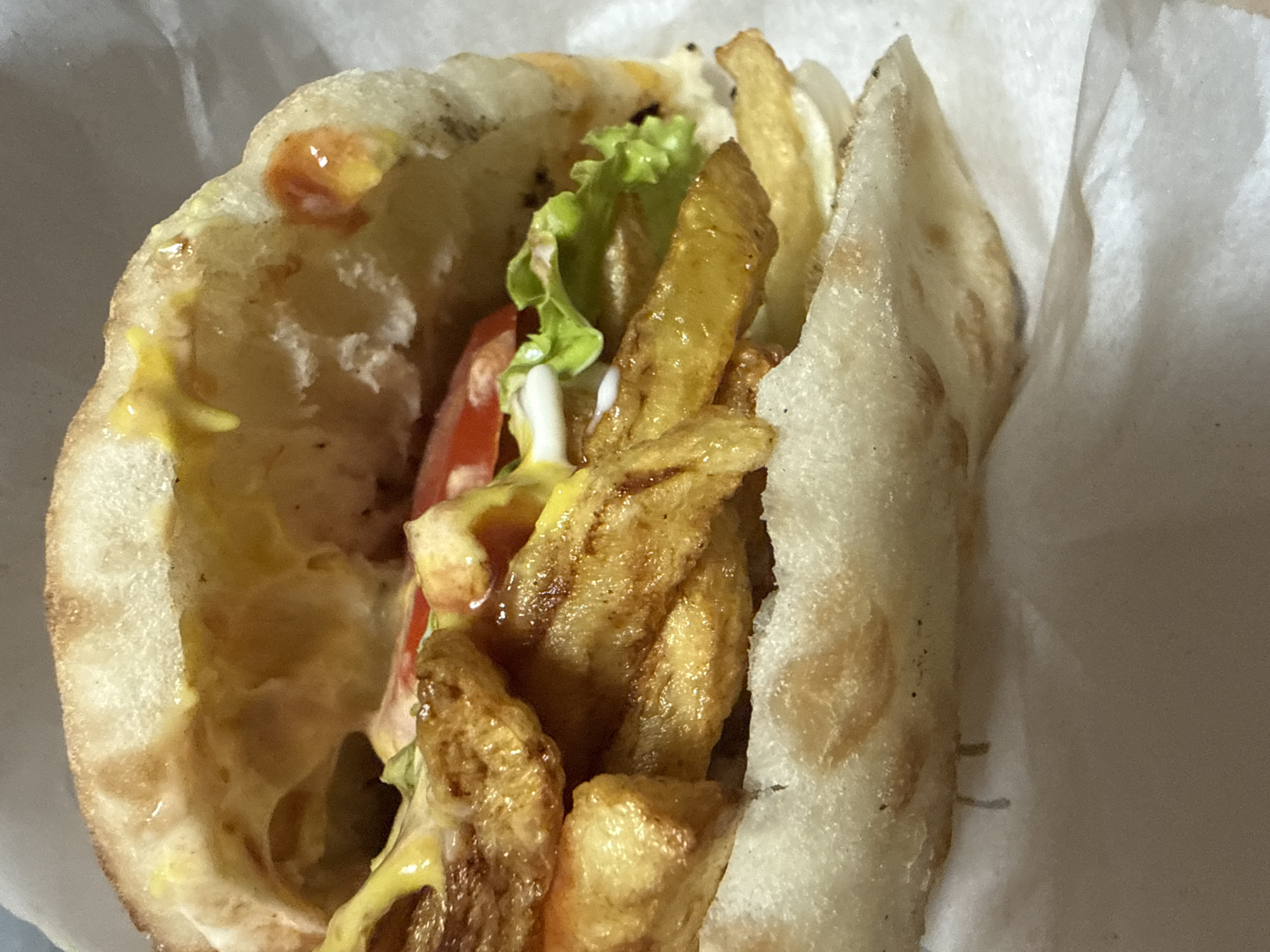 A close-up, slightly angled shot shows a sandwich filled with french fries, lettuce, tomato, and various sauces, including mustard and ketchup, resting on white paper. The sandwich bread is soft and slightly browned, with some areas appearing lighter than others. The french fries are golden brown and appear to be crispy. The lettuce is green and fresh, and the tomato slices are red. The sauces are visible as yellow and red drizzles and dollops. The background is mostly out of focus, with only a hint of white paper and a darker, textured surface. There are no people visible in the image. The sandwich appears to be a casual meal, possibly from a street food vendor or a fast-food restaurant. Due to the lack of identifiable landmarks or context within the image, it is not possible to determine the specific location within the city of Plovdiv, Bulgaria.