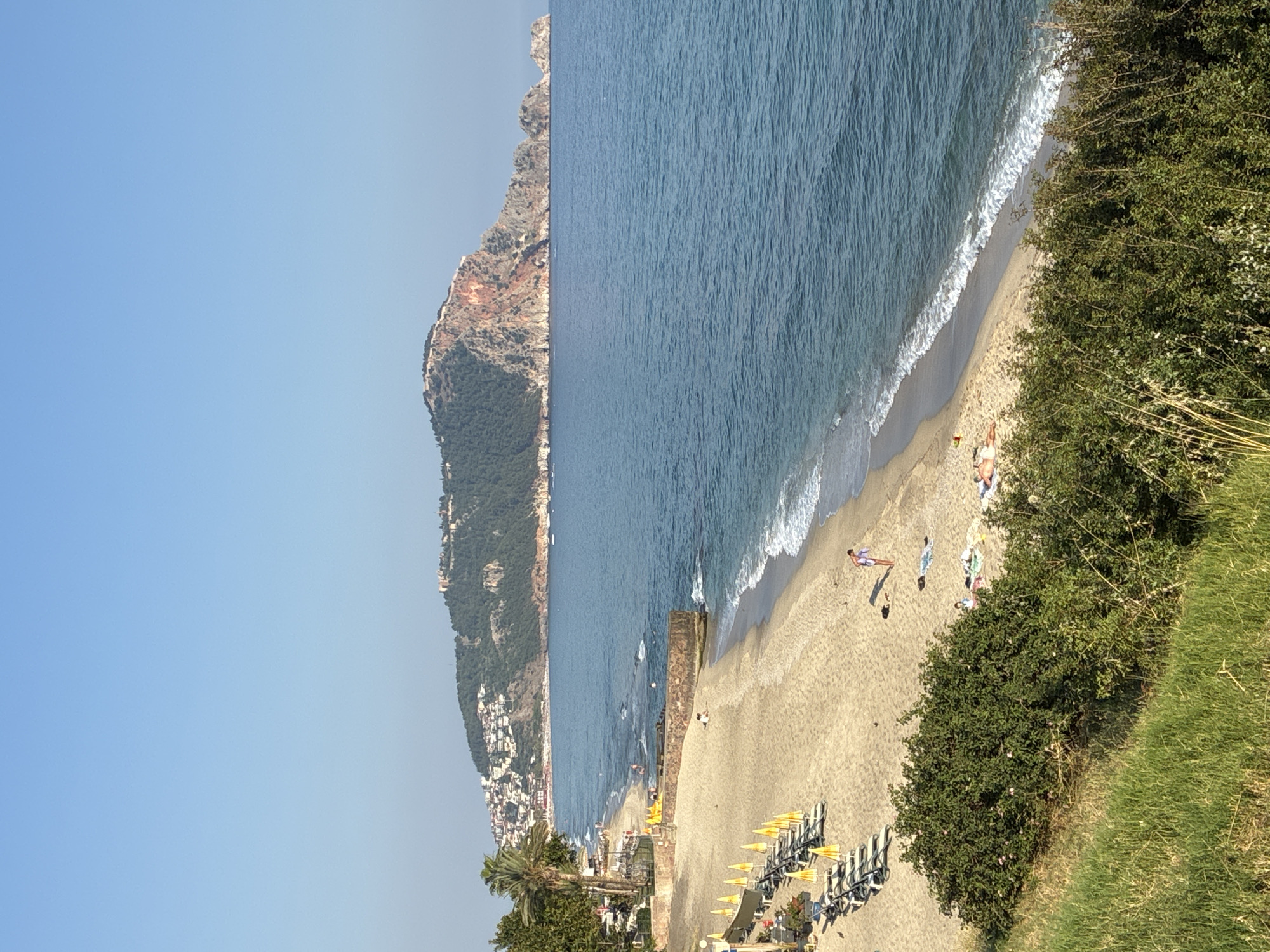 The image depicts a scenic view of a beach and the sea in Alanya, Turkey, with a prominent hillfort dominating the background. The foreground features a grassy embankment overlooking a sandy beach. On the beach, several rows of empty lounge chairs and yellow umbrellas are set up, suggesting a resort or public beach area. A few people are visible on the sand, including one individual standing near the water and others sunbathing. The sea is a deep blue, with gentle waves lapping at the shore. The background is dominated by a large, rocky hill covered in green vegetation and dotted with buildings, which is characteristic of the Alanya Peninsula and its famous castle. The sky is clear and blue, indicating a sunny day.