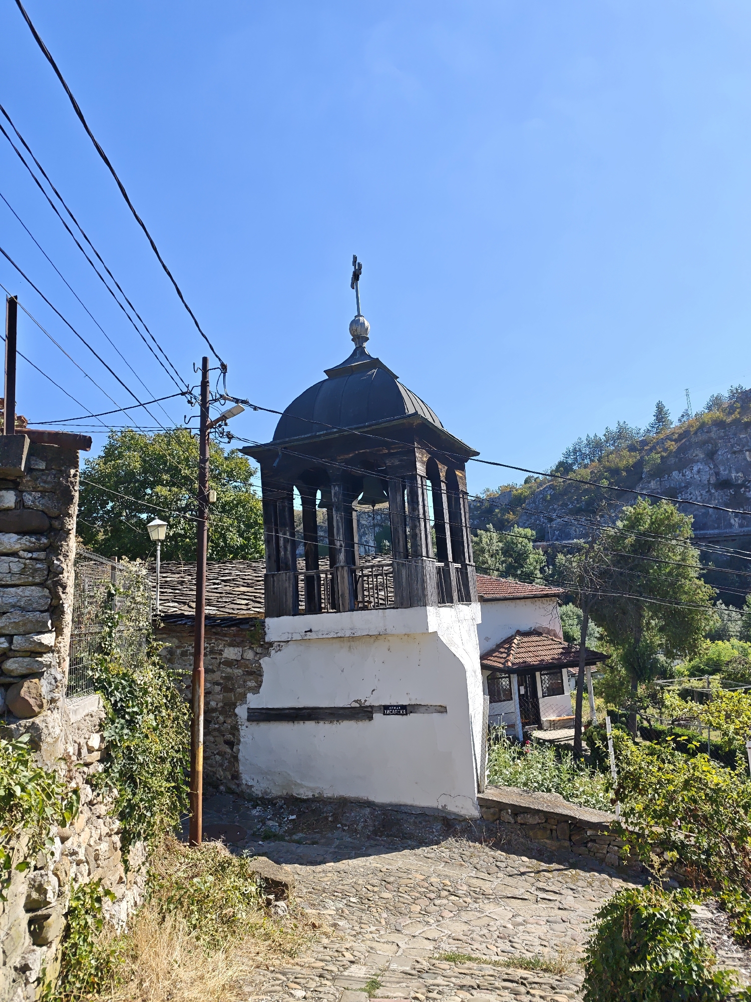 A bell tower with a dark domed roof topped with a cross stands prominently in the foreground of this image. The tower has a white base with some visible wear and tear, and an open, dark wooden upper section with a railing. The tower is situated on a cobblestone street. To the left of the tower, a stone wall covered in ivy and dry grass leads towards the viewer. Behind the bell tower, a building with a red tile roof and white walls is visible. The background features lush green trees and a rocky hillside under a clear blue sky. Power lines are strung across the upper part of the image, running over the bell tower and the surrounding buildings. The overall setting appears to be a historical village or town. This is the bell tower of the Saint George's Church in the Old Town of Lovech, Bulgaria.
