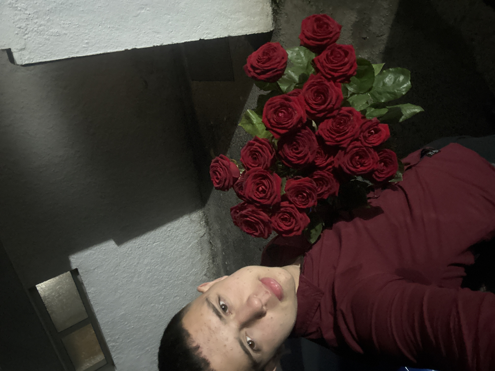 A young man is seen from a high angle, holding a bouquet of red roses. He is wearing a red shirt and appears to be outdoors, standing near a white, concrete wall. The background is dark, suggesting it might be night or a dimly lit area. The roses are clustered together and have visible green leaves. The location cannot be determined from the image.