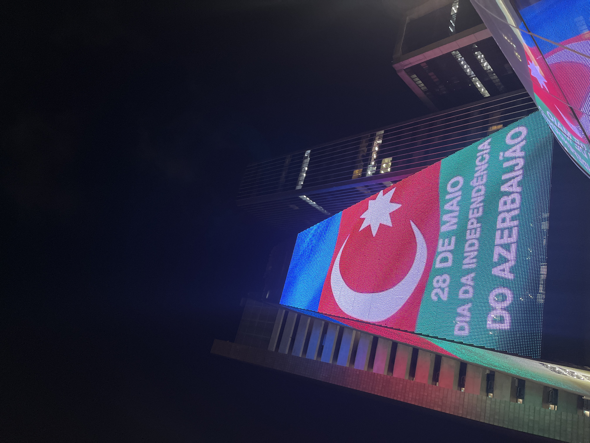 The image shows a low-angle view of a large LED display on the side of a building in an urban environment at night. The display features the flag of Azerbaijan, with horizontal bands of blue, red, and green, as well as a white crescent and eight-pointed star. Text in Portuguese reads "28 DE MAIO DIA DA INDEPENDÊNCIA DO AZERBAIJÃO," which translates to "May 28, Azerbaijan Independence Day." The buildings appear to be modern skyscrapers with illuminated windows. The location is unknown.
