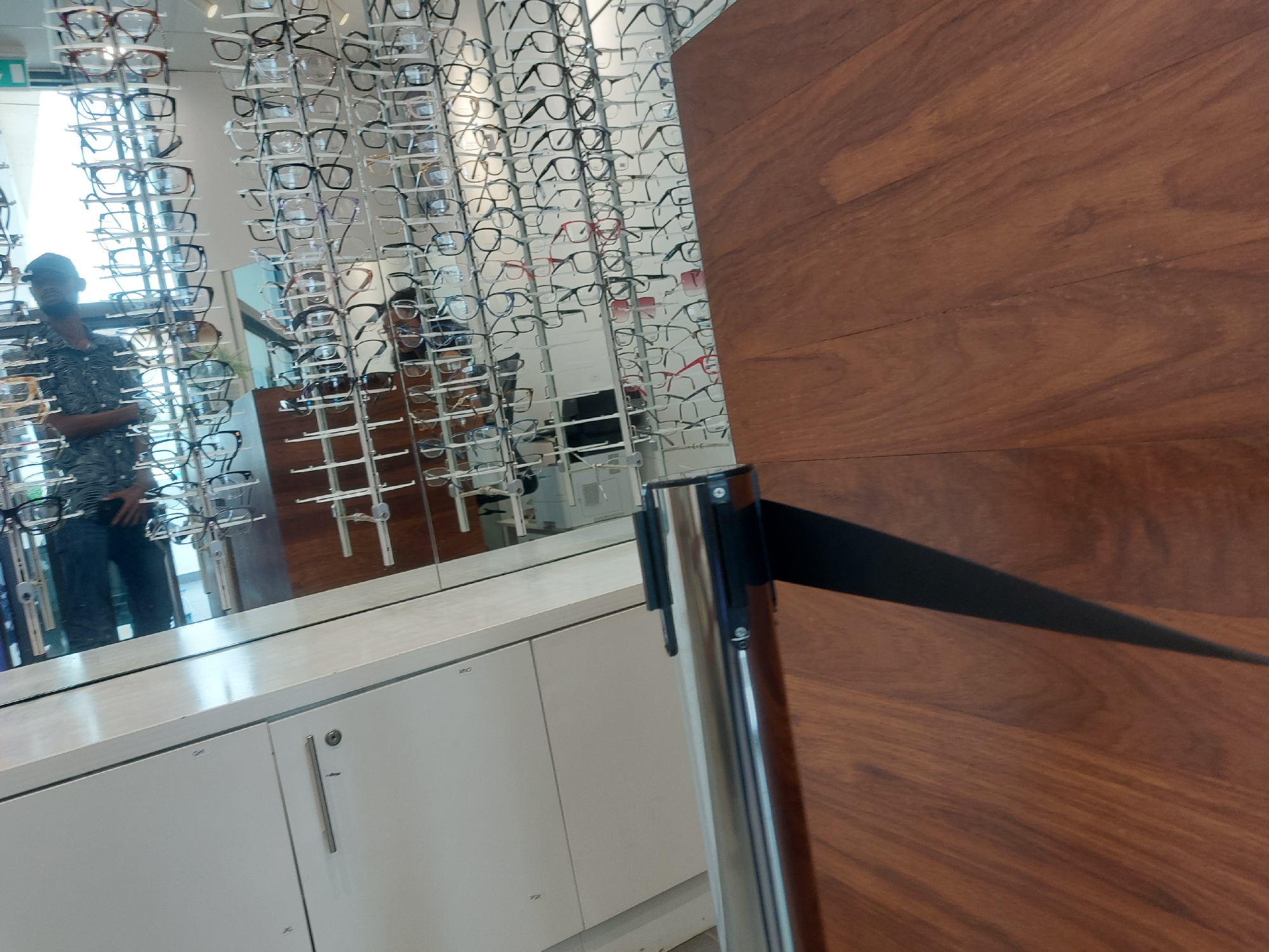 This indoor image captures the interior of an optometry store or an eyeglasses section, likely in Dar es-Salaam, Tanzania, with a strong focus on eyeglass displays and reflections.
The left side of the frame is dominated by a large mirror reflecting numerous eyeglasses frames displayed on vertical racks. A person is visible in the mirror's reflection on the far left, wearing a patterned shirt, jeans, and a baseball cap. They appear to be casually browsing the eyeglasses, with one hand near their waist. Further to the right in the reflection, another person is faintly visible, also seemingly engaging with the displays.
Below the expansive mirror, a sleek white counter or cabinet system extends horizontally. One section of the cabinet features a metallic handle and a keyhole. Faint alphanumeric labels like "VHMO," "SHI," "JKC," and "POJ" are barely visible on the white cabinet doors. In the top left corner of the reflected area, a green "EXIT" sign is discernible.
On the right side of the image, a tall, silver retractable belt barrier (stanchion) with a black, extended belt diagonally spans the space, directing flow or indicating a boundary. To its right, a warm-toned wooden wall or panel runs vertically, showcasing natural grain patterns. The overall scene suggests a modern retail environment dedicated to eyewear, with the reflected figures indicating customer presence.