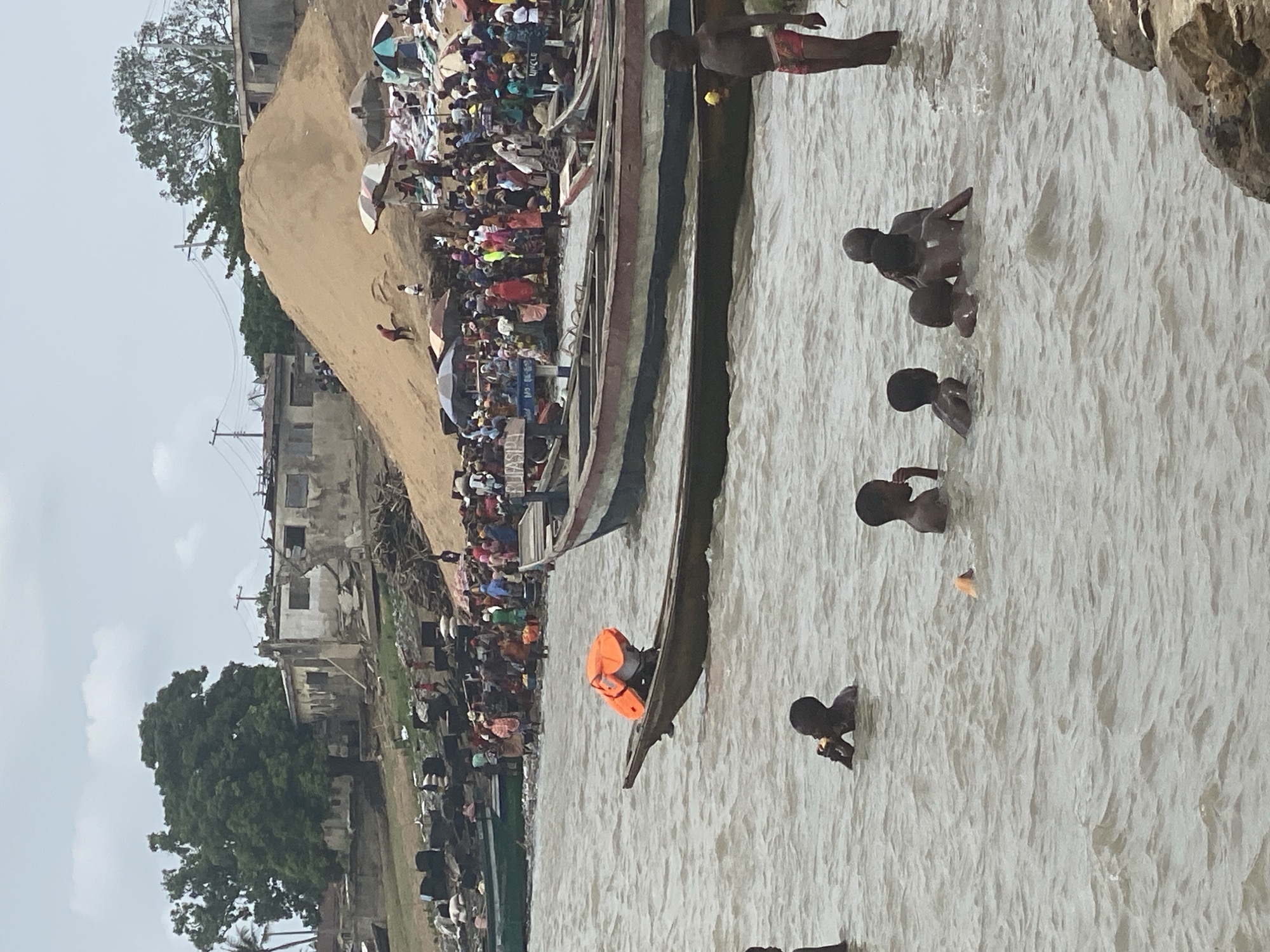 The image shows a crowded riverbank scene in Lokoja, Nigeria, with multiple people swimming in the murky water. Several wooden boats are docked along the shoreline, near a large gathering of people on a sandy bank. Buildings are visible in the background. At least seven children are swimming. One boat contains an orange life vest.