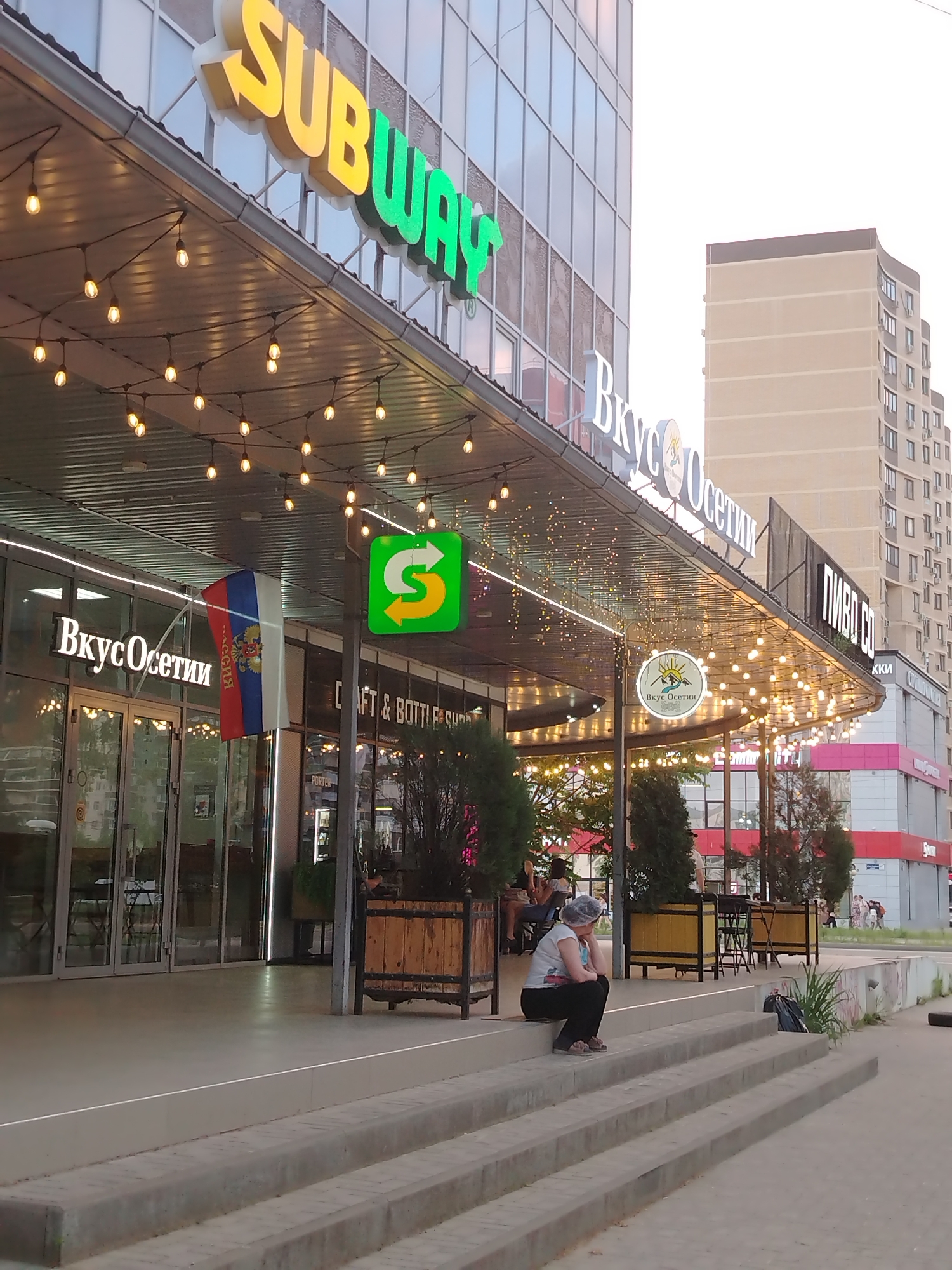The image shows the exterior of a Subway restaurant in Krasnodar, Russia. The restaurant is located on the ground floor of a modern building with a glass facade. The Subway logo is prominently displayed above the entrance. A Russian flag is hanging next to the entrance of a business identified as "ВкусОсетии" (Vkus Osetii), which translates to "Taste of Ossetia". String lights are hung under the overhang of the building. In front of the building, there are steps leading up to the entrance area. A person is sitting on the steps, facing away from the camera. In the background, other buildings and people are visible. The sky appears to be overcast, suggesting it might be evening or a cloudy day.