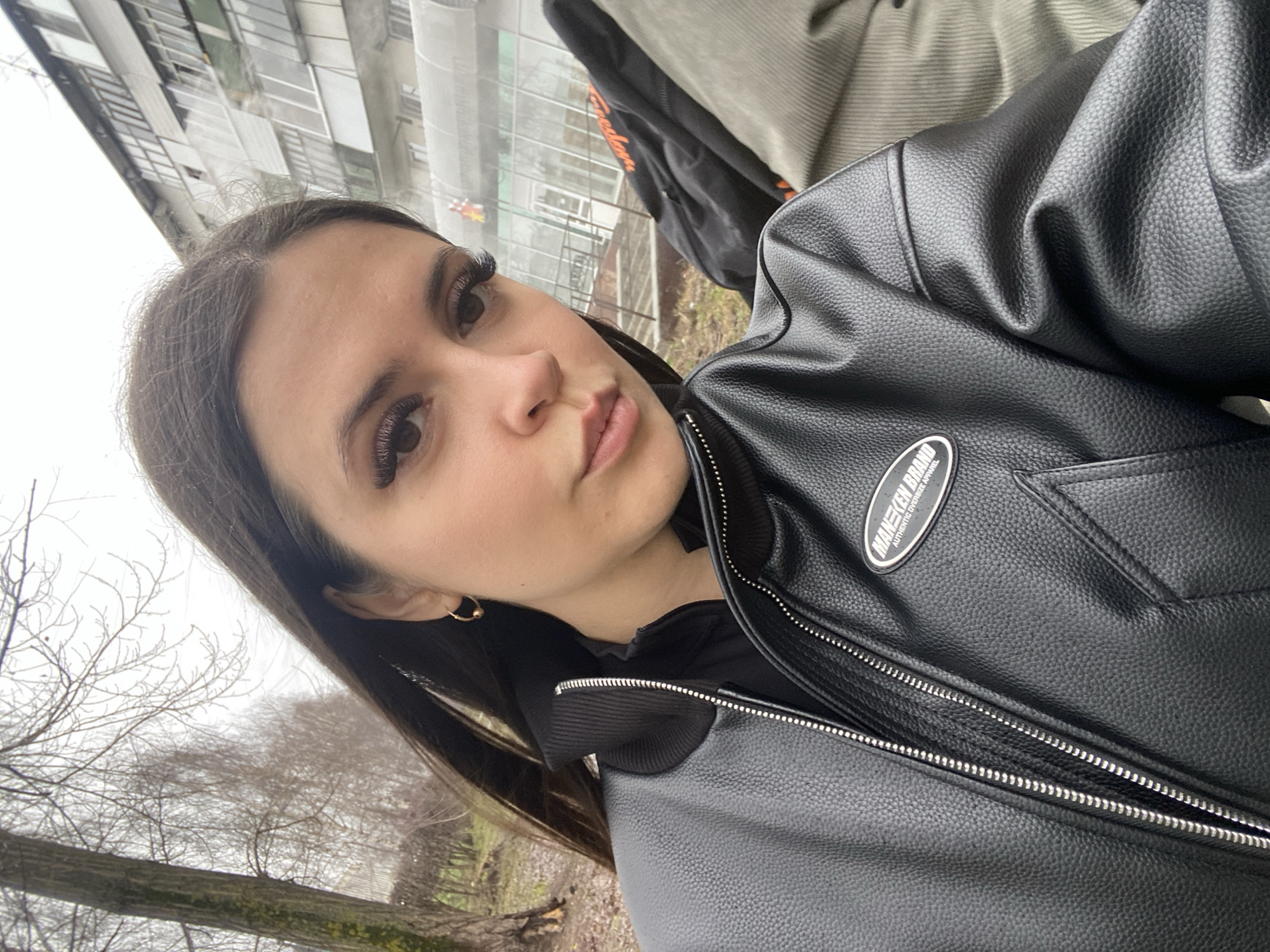A close-up, angled outdoor portrait features a young woman in Chaikovsky, Russia. Her dark hair frames her face, highlighting her prominent eyelashes and a neutral expression as she gazes slightly away from the camera. She wears a black faux leather jacket, featuring silver zippers and a patch on the left chest. The patch displays the text "MANEKEN BRAND" with "AUTHENTIC OVERSIZE APPAREL" beneath it. A small gold hoop earring is visible in her right ear. The background suggests an overcast daytime during a colder season, with bare trees and brown ground cover. Faintly visible in the distance are pale, multi-paned structures, resembling greenhouses or industrial buildings, under a dull, cloudy sky. An orange object or text is also barely discernible in the background.