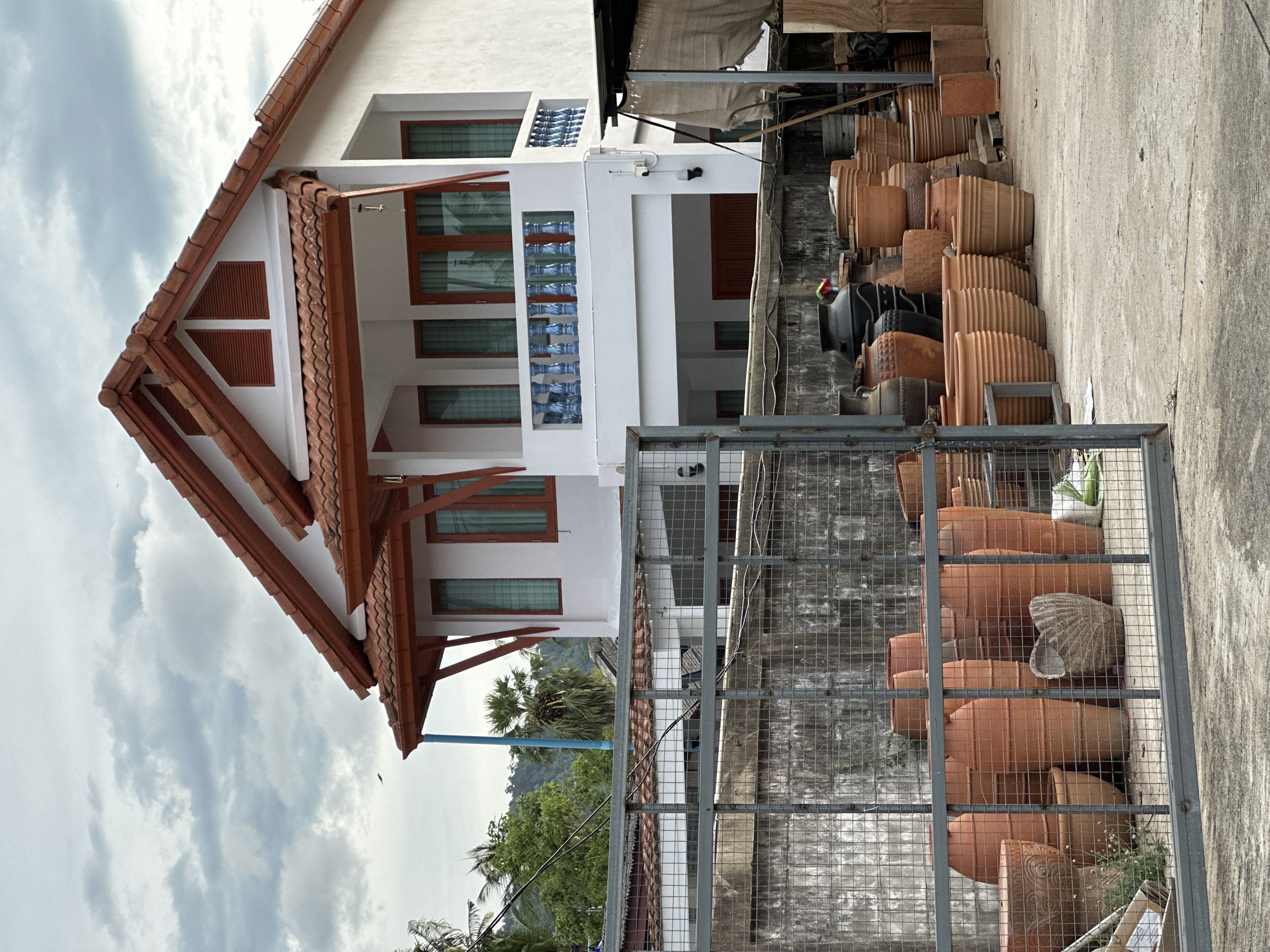 In Baan Mae Nam, Thailand, a white two-story house with a red-tiled roof is visible. The house features a second-story balcony with a blue and white railing and wooden-framed windows. In the foreground, a collection of large terracotta pots and planters are neatly arranged on a concrete surface, partially obscured by a grey metal fence. A concrete wall with a rough texture runs behind the fence and pots. The background includes lush green trees and hills under a cloudy sky.