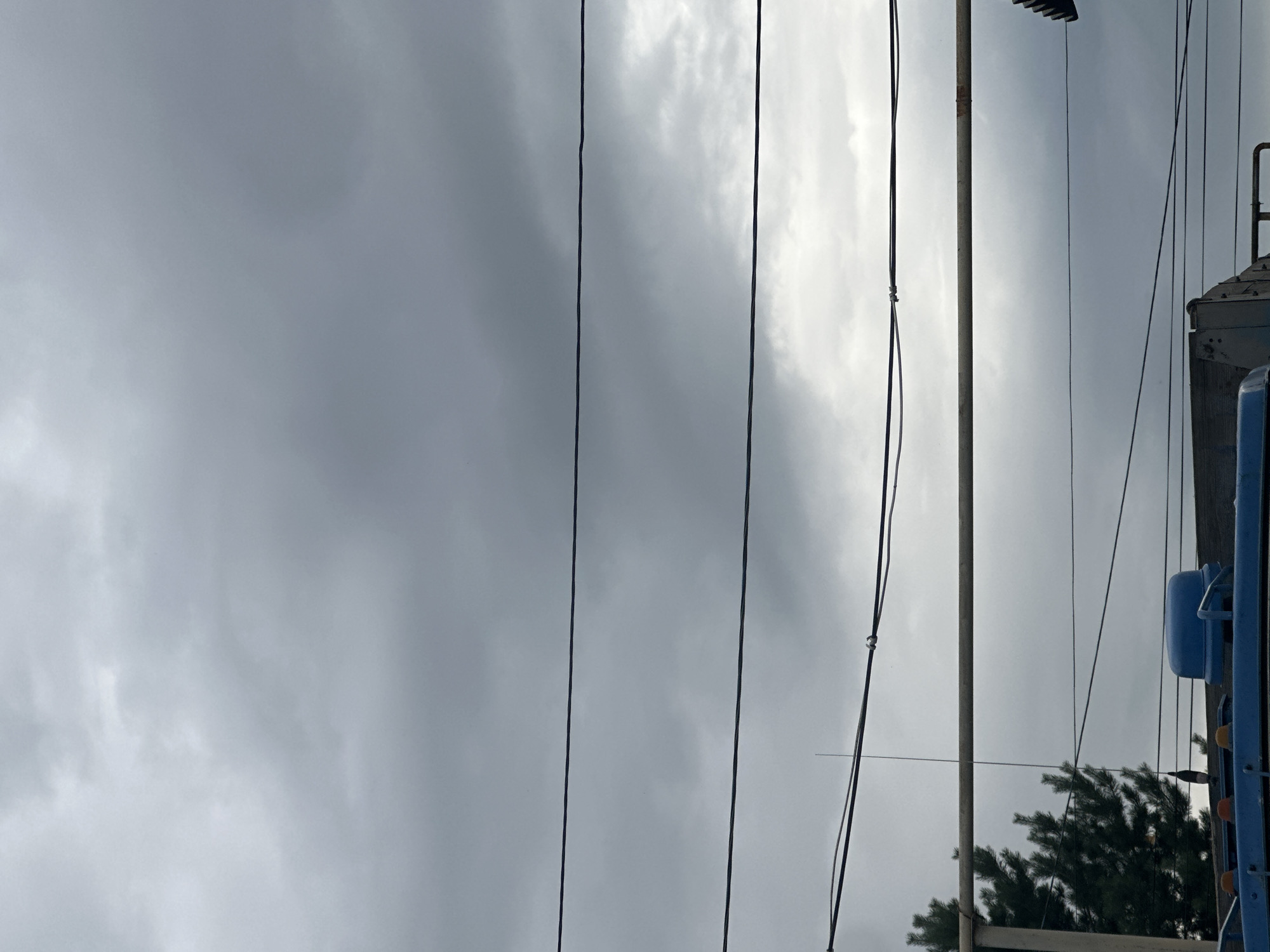 A cloudy sky fills the upper portion of the image, with distinct horizontal power lines crisscrossing the frame. In the lower section, a dark green coniferous tree is visible on the left, partially obscuring a tall, thin antenna that extends upwards. Below the antenna, a light-colored metal bar runs horizontally across the image. Further down, the blue top of a vehicle or structure is prominent, with several yellow and orange lights visible on its edge. The bottom of the image shows a portion of a blue and gray vehicle or structure, with a wooden edge on the left. The scene appears to be an outdoor setting in the city of Tobyl, Kazakhstan.