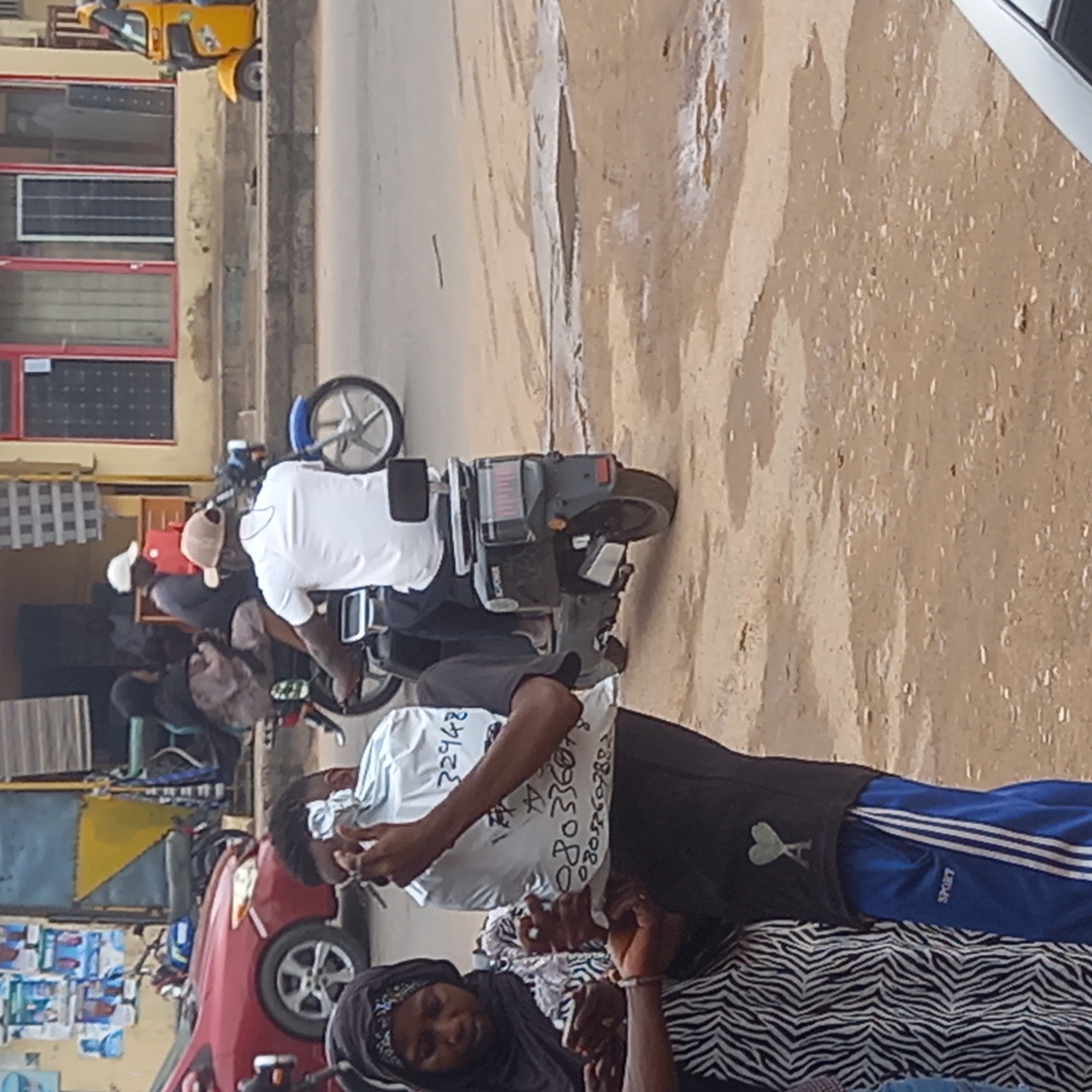 The image shows a street scene in Dadi Riba, Nigeria. Several individuals are present. A woman in a black hijab and zebra-striped garment sits near a man in blue track pants and a black shirt with a green logo. He is holding a white bag with text written on it. Another man is riding a black scooter, wearing a white shirt and tan hat. He appears to be interacting with someone on a nearby motorcycle. A red car is parked in the background, along with other motorcycles and a yellow tuk-tuk. The street appears to be paved and slightly dirty. Buildings with red and black windows are visible in the background.