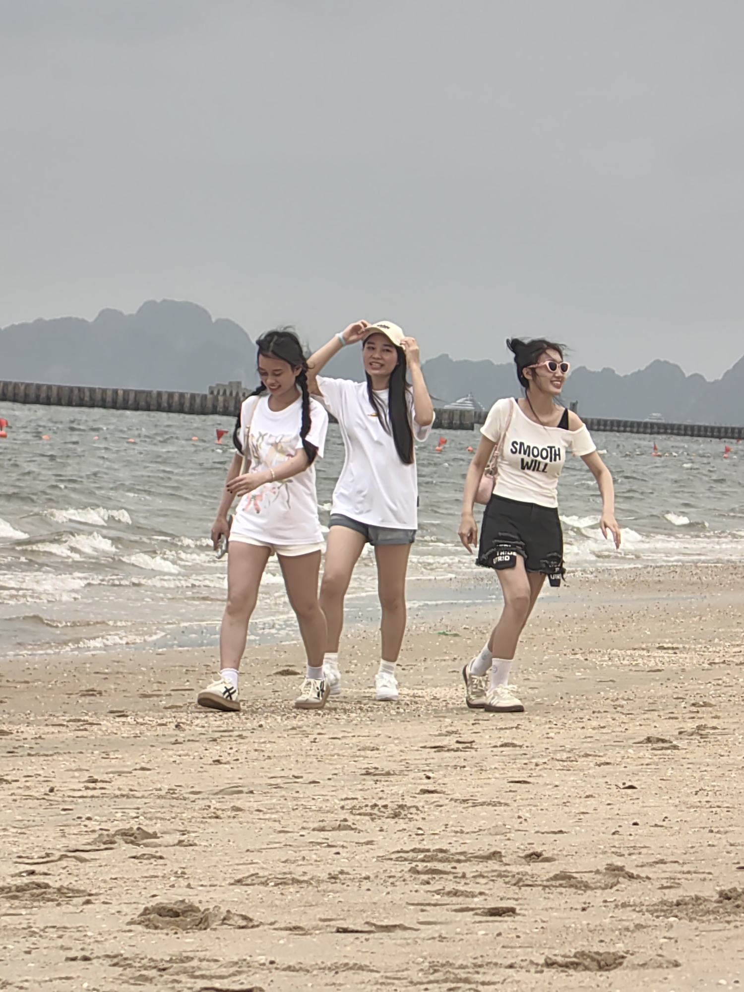 Three young women are walking on a sandy beach. The woman on the left has braided hair and wears a white t-shirt with floral designs and white shorts. The woman in the middle wears a white t-shirt, gray shorts, and a yellow baseball cap. The woman on the right wears sunglasses, a white t-shirt with the words "SMOOTH WILL" printed on it, and black shorts. A body of water laps at the shore behind them. A pier-like structure extends into the water, and mountains are visible in the background. The scene is overcast. The location is Bai Chay Ward, Vietnam.