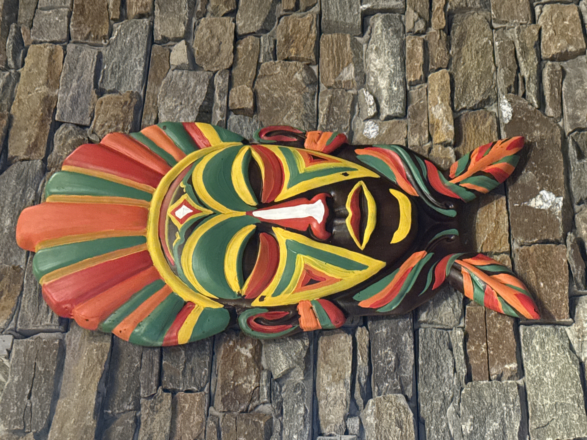 A vibrantly colored, carved wooden mask hangs on a stone wall. The mask is dark brown with a headdress of red, green, and orange fan-like structures. The face is painted with bold geometric patterns in yellow, green, red, and white, featuring large eyes, a white nose, and red lips. Two leaf-like appendages with similar coloration hang from the bottom sides of the mask. The background is a textured wall made of irregular, grey and brown stones. There are no people or actions depicted in the image. The location is Wuxing District, China.
