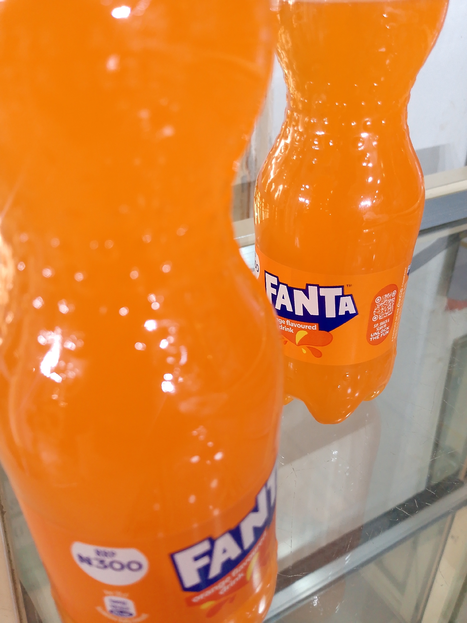 Two orange plastic bottles of Fanta, a carbonated soft drink, are displayed on a glass surface. The bottle in the foreground is out of focus, while the bottle in the background is in focus and shows its label. The label prominently features the Fanta logo in blue and white, along with text indicating it is an "Orange flavoured drink." A QR code and instructions to "SIP, SNACK & SCAN TO UNLOCK THE FUN" are also visible on the label. A price tag reading "N300" is present on the front of the foreground bottle. The background is a lightly colored, indeterminate wall. The setting appears to be a retail environment, potentially a shop in Bukuru, Nigeria, given the currency notation.