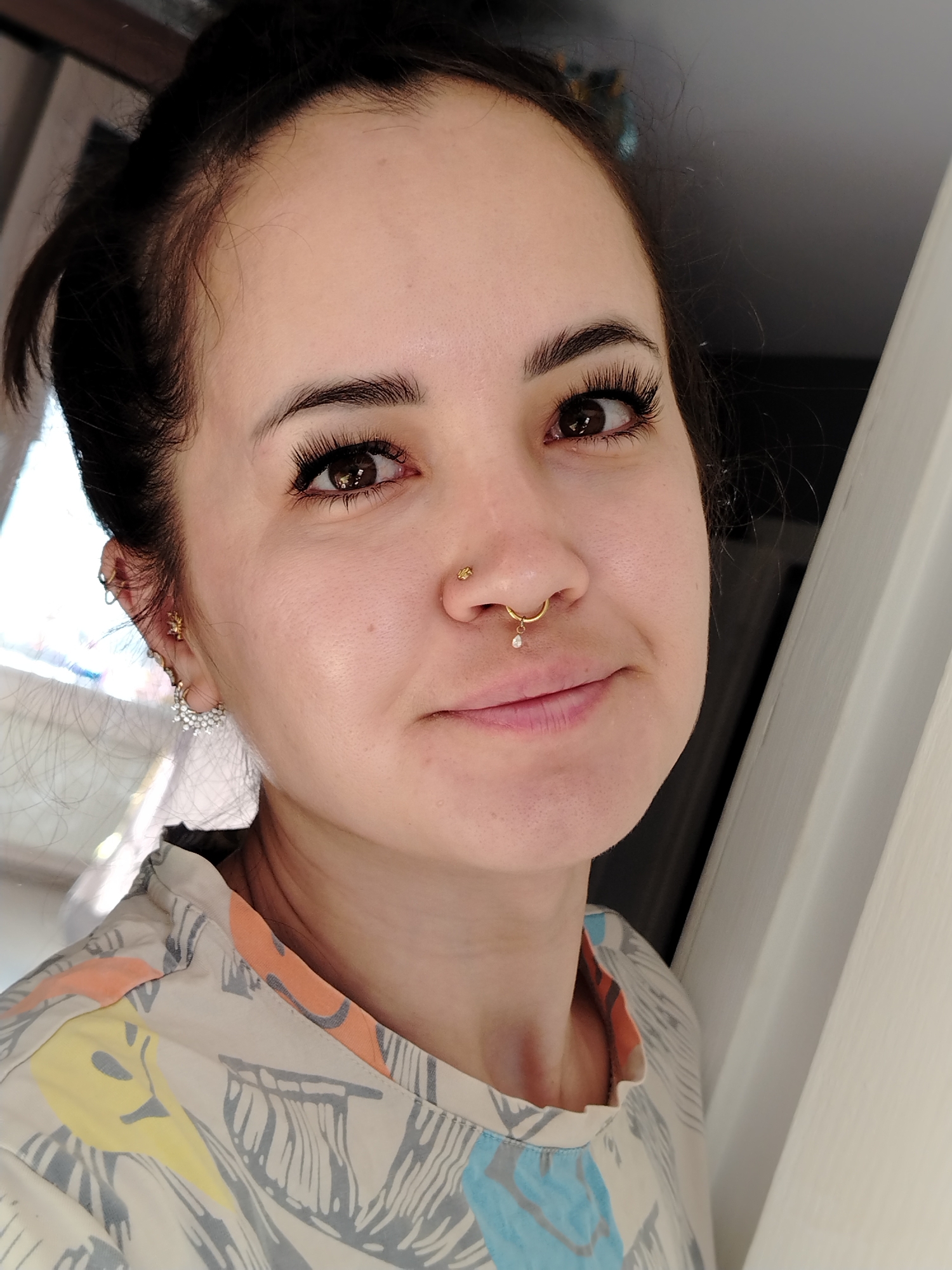 A close-up, shoulder-up, medium shot captures a young woman with dark hair pulled back and a septum piercing featuring a gold ring with a small diamond charm. She also has multiple earrings in her left ear, including a hoop with small diamonds. Her makeup includes long, dramatic false eyelashes and subtle foundation. She is wearing a light-colored, collared shirt with a pattern of stylized gray leaves and colorful abstract shapes in yellow, orange, and blue. The background is out of focus, with hints of natural light coming from the left and a plain white door or wall to the right. The overall impression is a casual, indoor portrait.