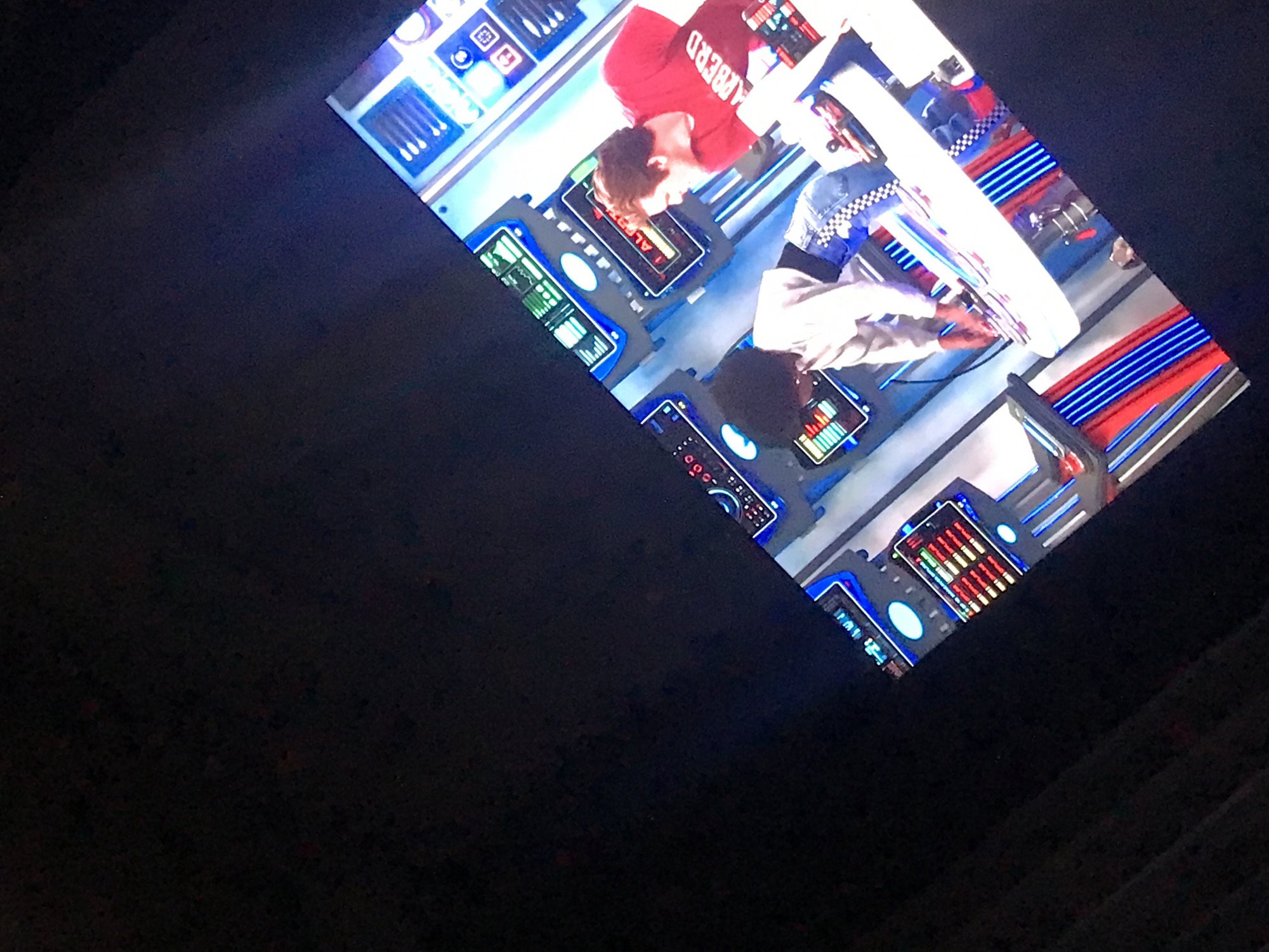 The image shows a television screen displaying two people in what appears to be a sci-fi setting, possibly a cockpit. One person is wearing a red shirt with the word "BARBERD" printed on it, while the other is wearing a light-colored jacket with a checkered collar. They seem to be operating some kind of control panel with various lights, buttons, and monitors. The setting has blue and red striped accents. The television is located indoors with a dark background surrounding it. There is no information to suggest this image was taken in Yerevan, Armenia.