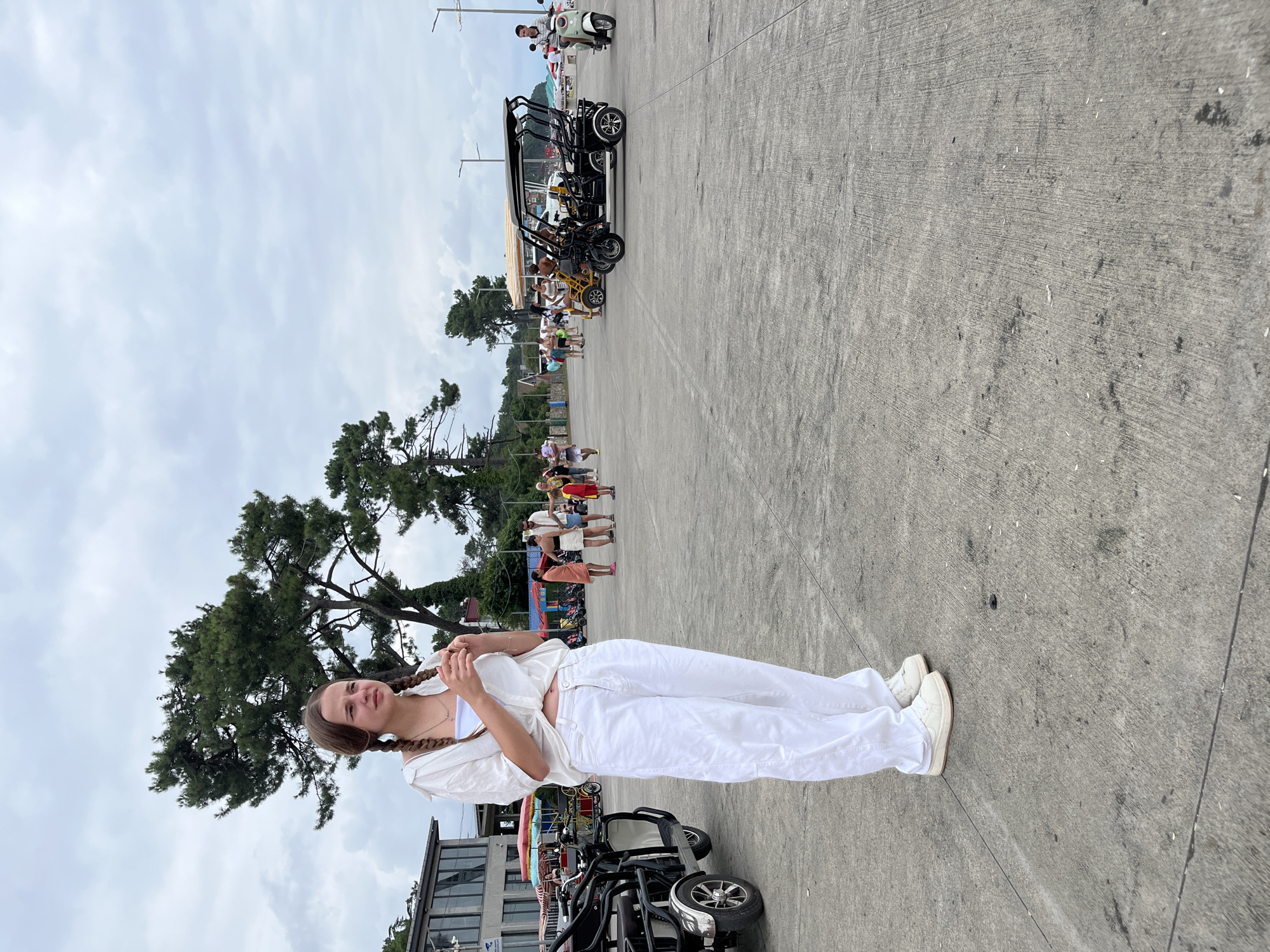 A young Caucasian woman with braided brown hair stands in the foreground of an outdoor scene in Ureki, Georgia. She is wearing a white open button-up shirt, a white bandeau top, and loose-fitting white pants, along with white sneakers. Her arms are slightly bent, and her hands are near her chest. The background reveals a cloudy sky and a paved area with several people and vehicles. To the left, a three-wheeled electric vehicle is parked. In the middle ground, a group of people are visible, some walking and others standing near brightly colored tents. Further back, more people and parked vehicles, including golf cart-like vehicles and a scooter, are present. Trees line the left side of the background, and a body of water can be seen in the distance. The overall setting appears to be a promenade or public square near a beach or resort area.