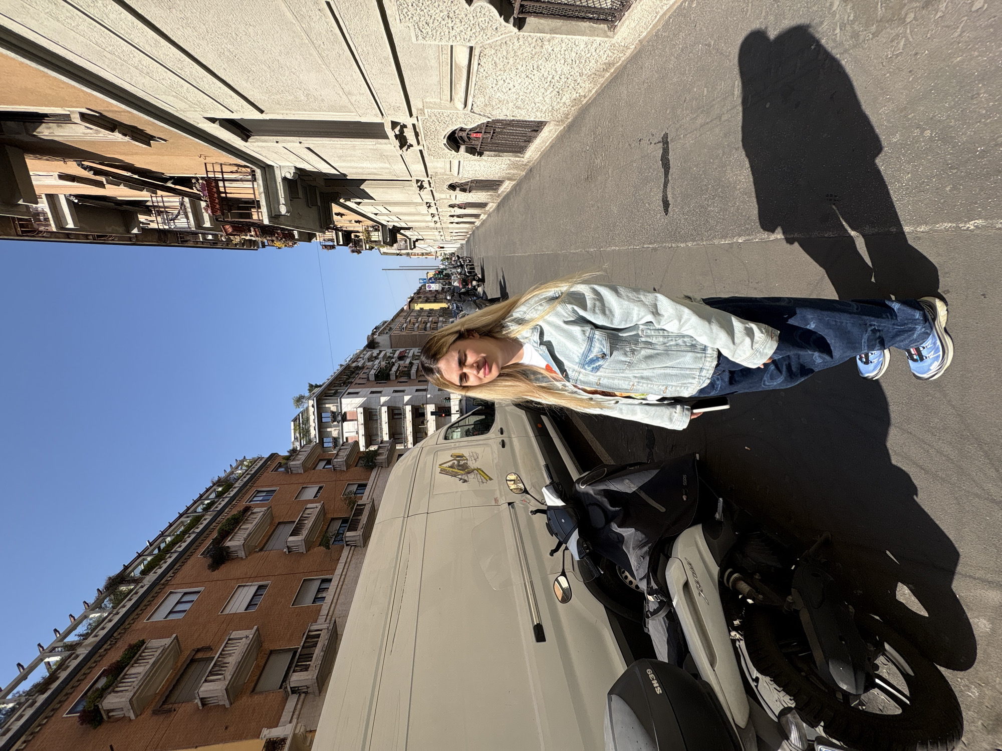The image captures a street scene in Milan, Italy, characterized by tall buildings with balconies on both sides, casting long shadows on the street due to the angle of the sun. A young woman with long blonde hair is standing on the street, wearing a denim jacket and jeans, and light blue sneakers. To her right, a white van and a scooter are parked. The overall atmosphere suggests a sunny day in an urban environment.