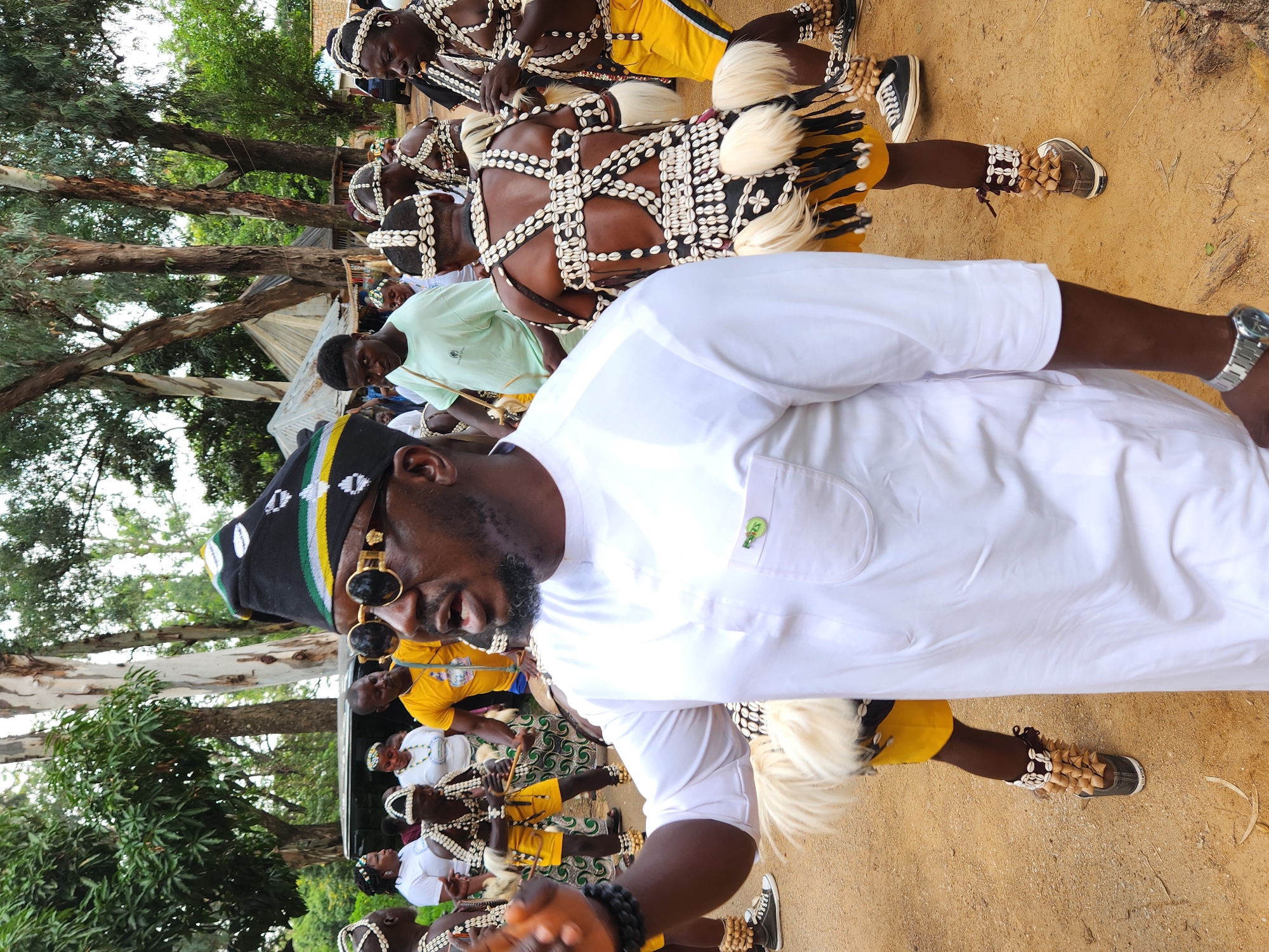 This is a medium shot, eye-level, outdoor photograph taken in the city of Jos, Nigeria, during what appears to be a cultural event or festival. The foreground is dominated by a man in a white traditional outfit, a black and green cap, and gold-rimmed sunglasses. He is facing left and speaking, with his mouth open. His attire includes a white tunic with a breast pocket that has a small, green, circular emblem. He wears a silver wristwatch on his left wrist and a black beaded bracelet on his right.
Behind him, several other individuals are visible, many adorned in traditional ceremonial wear. These outfits consist of dark fabric, embellished with numerous white cowrie shells and adorned with white fluffy accents, possibly made of animal fur. Some of these individuals wear headdresses and have shell embellishments around their necks and arms. One person in the mid-ground wears a bright yellow t-shirt and is looking towards the left. Another person in a light green t-shirt is looking directly at the camera.
The background features tall trees with sparse foliage, indicative of an outdoor, possibly semi-arid environment, consistent with parts of Jos. Some tents or temporary structures are also visible in the background, suggesting a gathering or performance space. The ground is covered in light brown, sandy soil. The lighting suggests it is daytime, with diffused light possibly indicating an overcast sky or shaded area. The overall atmosphere appears festive and communal.