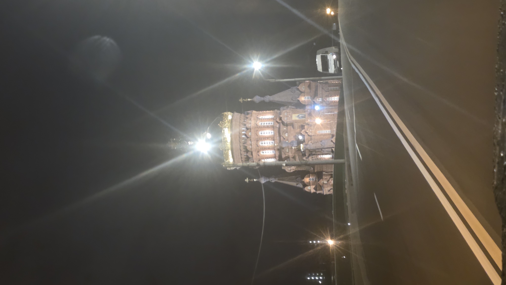 A night scene depicts an elaborate architectural structure illuminated by bright streetlights, juxtaposed against a dark sky and a portion of a roadway.
The building, which appears to be an elaborate temple or cathedral, is the focal point. Its design includes multiple tiers, detailed ornamentation, and a prominent golden dome. The façade is adorned with small windows and decorative elements, lit to accentuate its intricate design.
The setting is dark, with the sky appearing black due to the nighttime conditions. Streetlights emit strong beams of light, creating bright spots and long, contrasting shadows. A portion of a multi-lane roadway is visible, marked with white lane dividers that run diagonally across the frame.
The location is Saint Petersburg, Russia.
