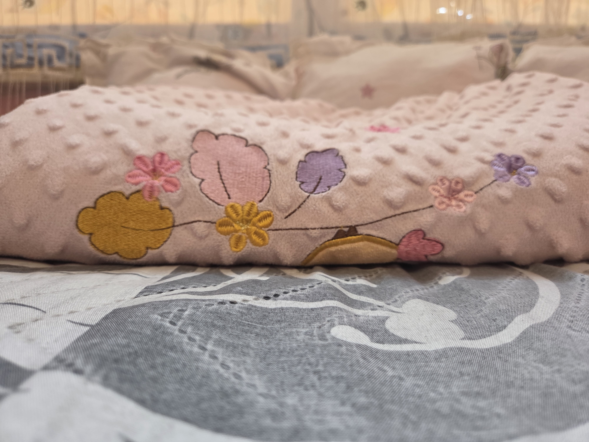 The image provides a close-up, low-angle perspective of bedding in an indoor setting, likely a bedroom in Krastevich, Bulgaria. A prominent light pink pillow or cushion lies horizontally in the mid-ground. Its surface is textured with numerous small, raised dots, giving it a soft, tactile appearance. Embroidered onto the side of the pillow facing the viewer are stylized floral and cloud-like designs in various colors. These include larger cloud-like shapes in light pink and lavender, and a bright yellow one, all connected by thin dark lines that resemble stems. Smaller, distinct flowers are embroidered in bright pink and yellow hues.
Beneath the pink pillow, a bedsheet is visible, featuring a pattern composed of different shades of grey, white, and black, with abstract or geometric shapes. The sheet shows subtle folds, suggesting it is in use. The background is softly blurred, revealing additional light-colored pillows or bedding, and a diffused light source, possibly a window with sheer curtains, creating a gentle illumination. The soft lighting suggests either early morning or late afternoon. No activity or event is taking place, and no individuals are present. The scene conveys a sense of comfort, domesticity, and tranquility. There is no visible text within the media file.