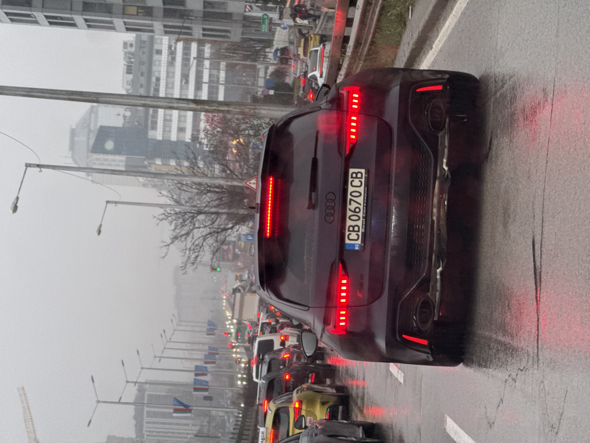 The image shows a traffic jam on a rainy day in Sofia, Bulgaria. The focus is on the rear of a dark-colored Audi RS3, identifiable by its distinctive tail lights, rear diffuser, and Bulgarian license plate "CB 0670 CB". The surrounding traffic includes a variety of cars, such as a yellow car, which are visible in the queue. Streetlights and tall buildings line the road, indicating an urban setting. Along the side of the road there are also flag poles with flags that appear to be Bulgarian. The weather is overcast, and the pavement is wet, reflecting the taillights of the vehicles. Overall the weather conditions appear to be poor with visibility limited to medium ranges.