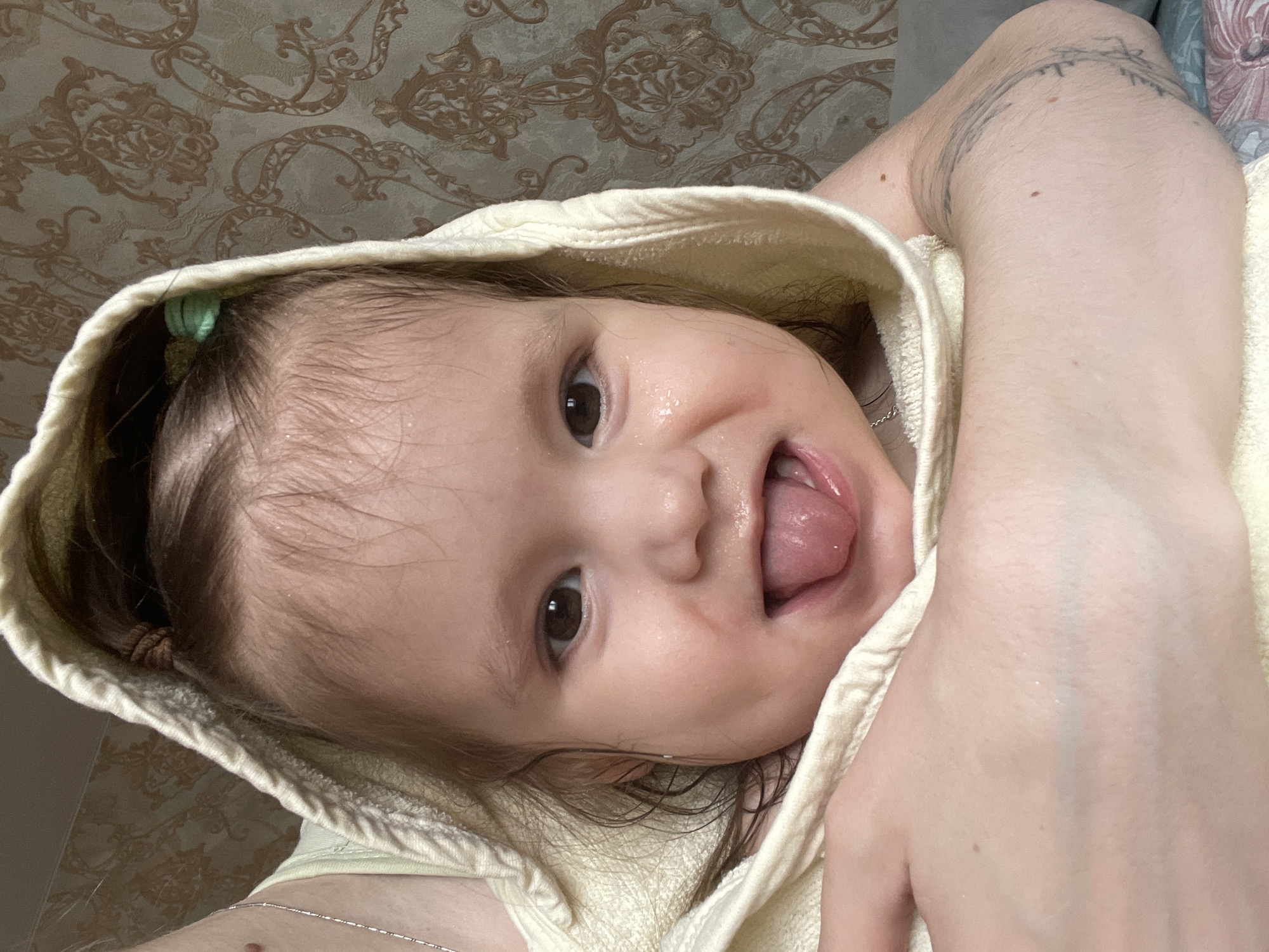 The image, taken indoors in Izhevsk, Russia, captures a playful moment between a baby and an adult. A young baby or toddler is the primary subject, wrapped in a light yellow or cream-colored hooded towel or garment, suggesting a recent bath due to its wet hair. A small, light-green hair tie is visible in the baby's hair on one side, with another subtle hair tie or bun on the opposite side. The baby looks directly into the camera with wide, dark eyes, displaying a happy and mischievous expression by sticking out its tongue, with visible wetness around its mouth and cheeks.
The adult holding the baby is partially visible. A fair-skinned arm and shoulder with a dark, intricate tattoo (appearing to be script or decorative patterns) are seen on the right side of the frame. Another hand, also fair-skinned, is visible at the bottom. A thin silver chain necklace is draped around the adult's neck/chest area. The background consists of a wall covered in patterned wallpaper featuring an ornate, damask-like design in shades of brown and beige. The image conveys a sense of warmth, care, and playful affection between the subjects. There is no visible text.