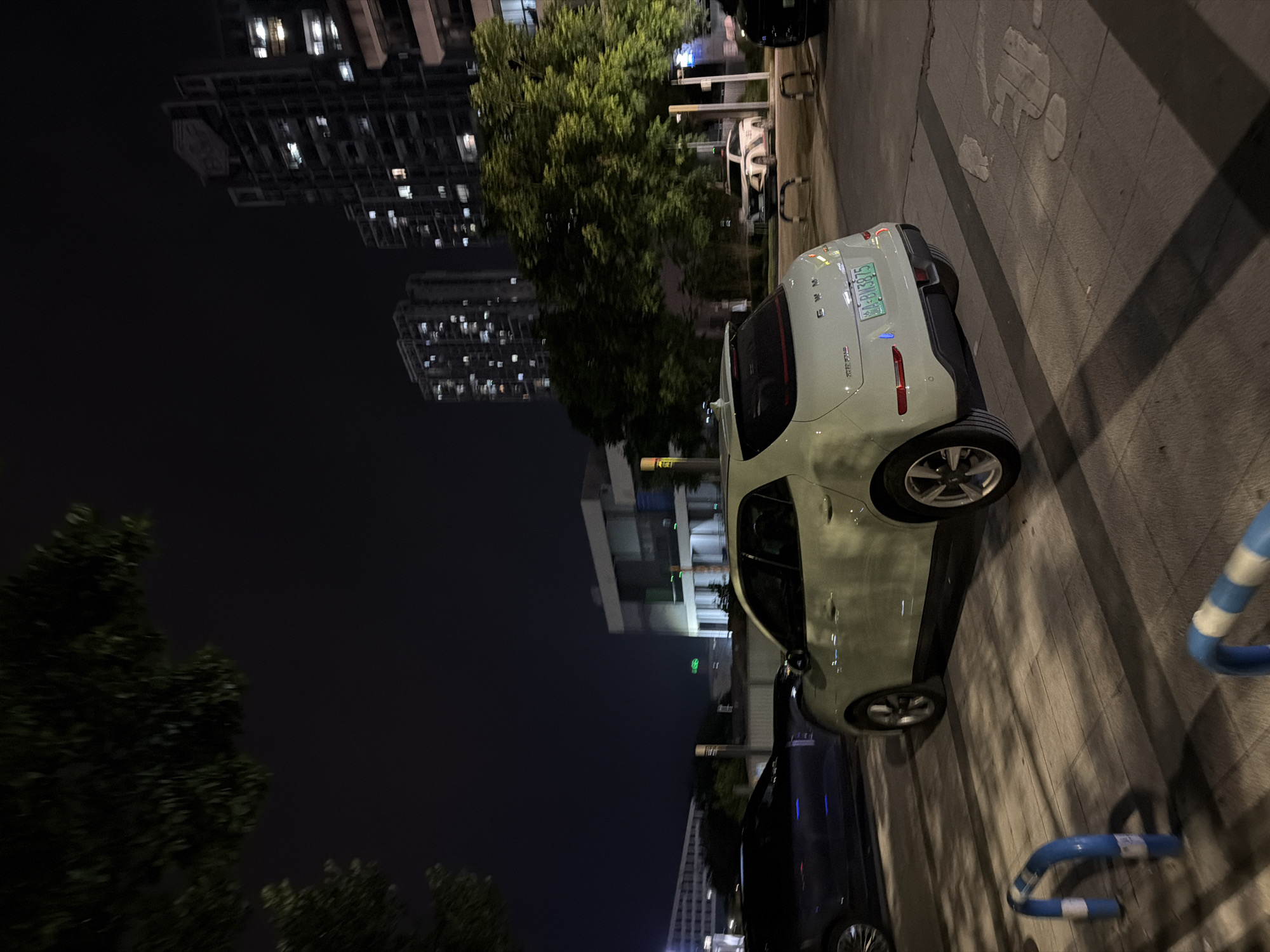 The image depicts a nighttime scene on a paved street in the Chongqing High-tech Zone, China. A light green hatchback car is parked, facing away from the camera, on the right side of the frame. The car's license plate is visible, reading "60-BW3875". To the left of the hatchback, a dark-colored sedan is partially visible. In the background, tall apartment buildings with illuminated windows are prominent against the dark night sky, indicating an urban setting. Trees with dense foliage are present on the left and in the mid-ground, casting shadows on the ground. The pavement in the foreground features blue and white striped bollards, suggesting a pedestrian or parking area. The overall environment appears to be a well-lit urban street or parking lot at night.