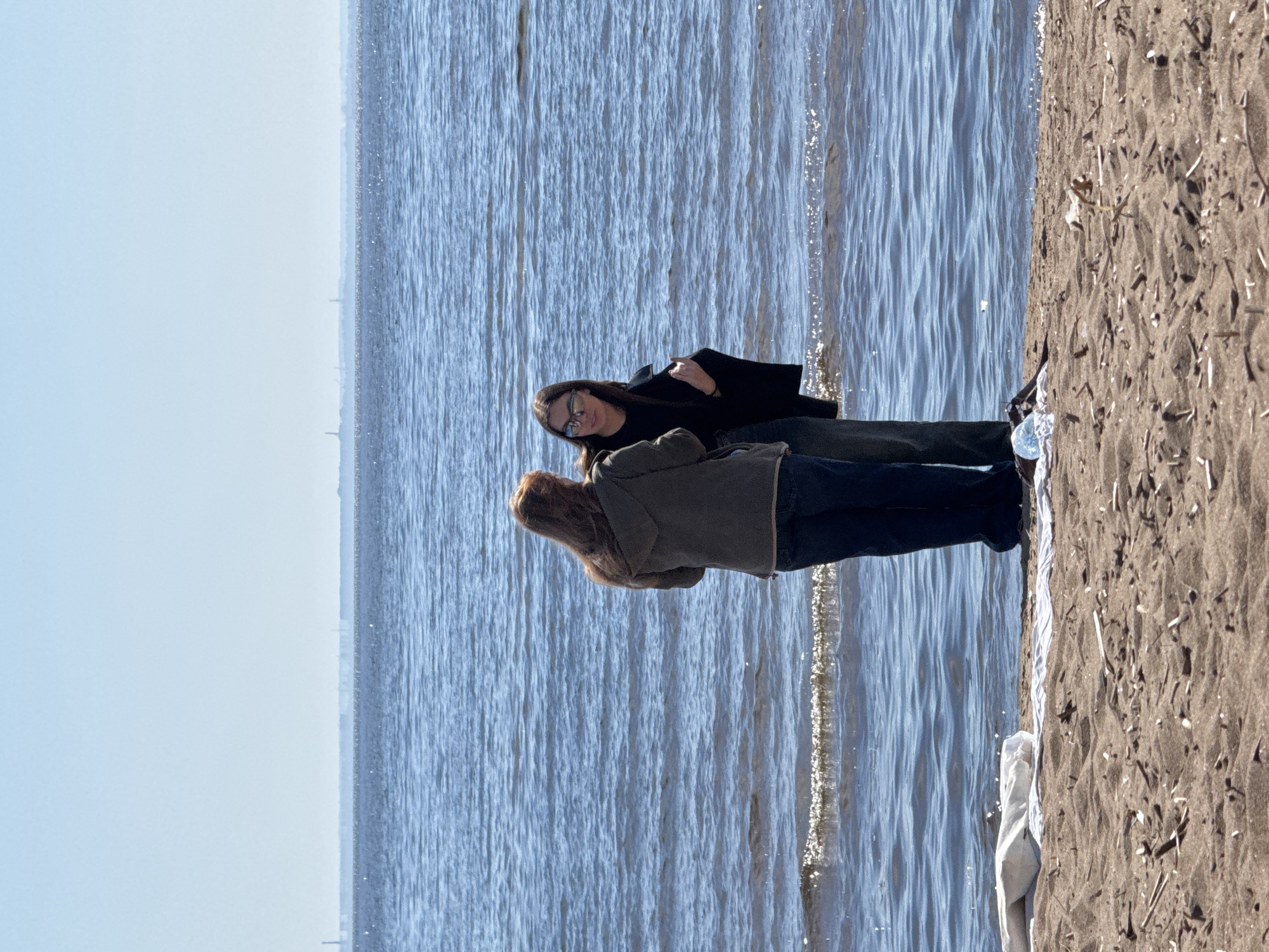 Two women are standing on a beach in Komarovo, Russia, near the water's edge. One woman has long dark hair and is wearing a black coat and sunglasses. The other woman has light brown hair and is wearing a green jacket. They are looking at each other. The beach is sandy, and the water is blue. The sky is light blue.