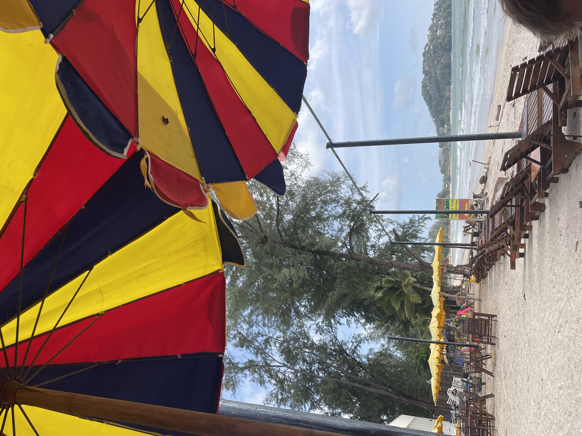 The scene depicts a beach in Choeng Thale, Thailand. Foreground: The undersides of two beach umbrellas, colored yellow, red, and blue, dominate the view. Midground: Beach chairs and small tables are lined up along the beach. Some are shaded by yellow beach umbrellas. Trees are interspersed between the umbrellas. Background: The ocean stretches out, leading to a landmass in the distance. The sky is partly cloudy.