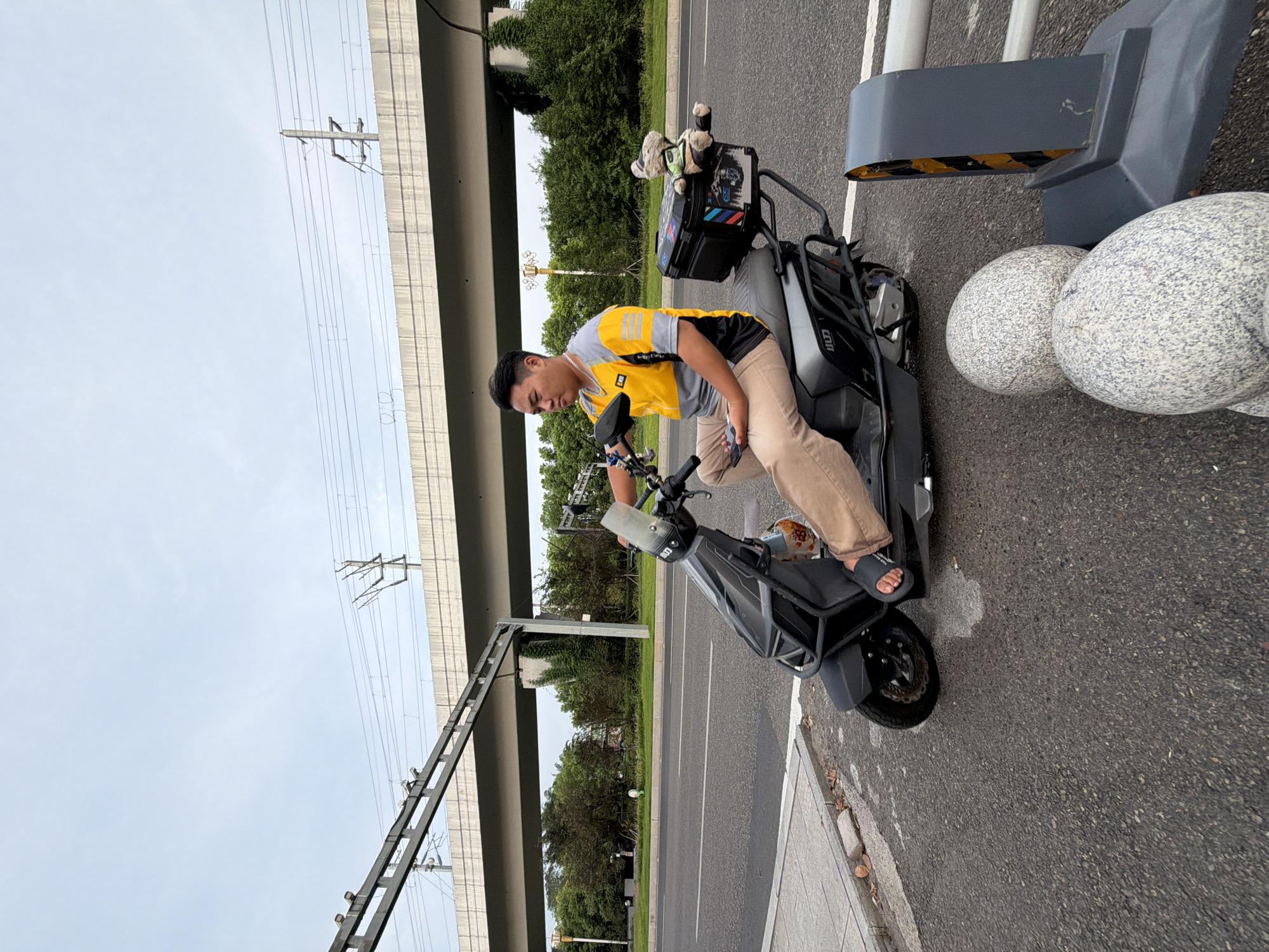 An Asian male wearing a yellow and gray shirt and tan pants is seated on a dark gray electric scooter on an asphalt road. The scooter has a black storage box on the rear, adorned with stickers, and a plush toy sitting on top of the box. The man holds a smartphone in his left hand. In the background, a concrete overpass with overhead power lines is visible, with lush green trees and foliage beyond. The scene appears to be on a road in Jiangjin, China, possibly near an elevated transportation route. The lighting suggests it is daytime, with an overcast sky.