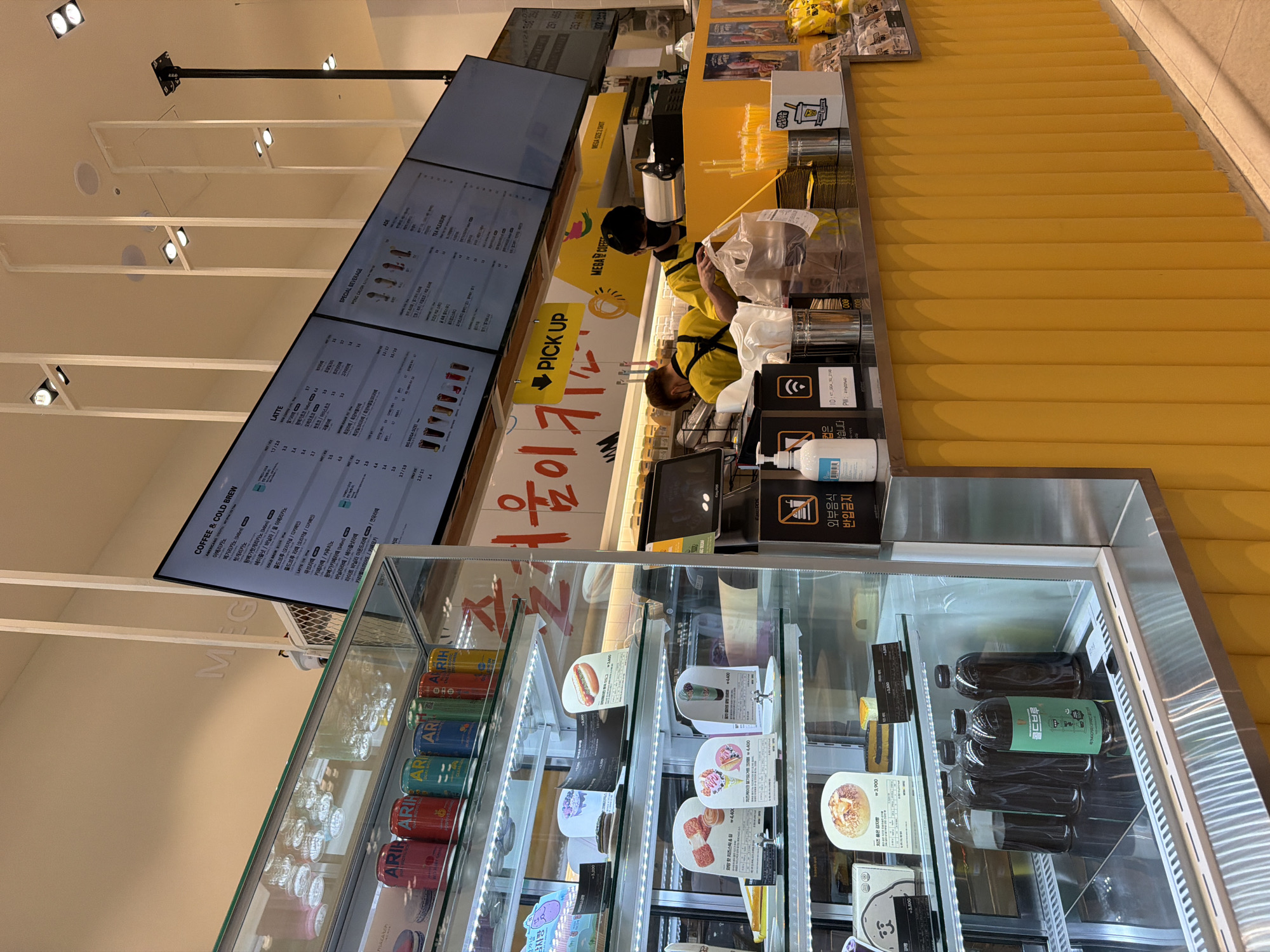 The image shows the interior of a Mega Coffee shop in Seoul, South Korea. The shop features a bright yellow counter with a menu displayed on screens above. Two employees, wearing yellow shirts and black hats, are working behind the counter, one of whom is interacting with a customer's order. Various food and drink items are displayed in a refrigerated glass case to the left, including canned beverages, cakes, and pastries. The overall environment is clean and modern, with ample lighting.
