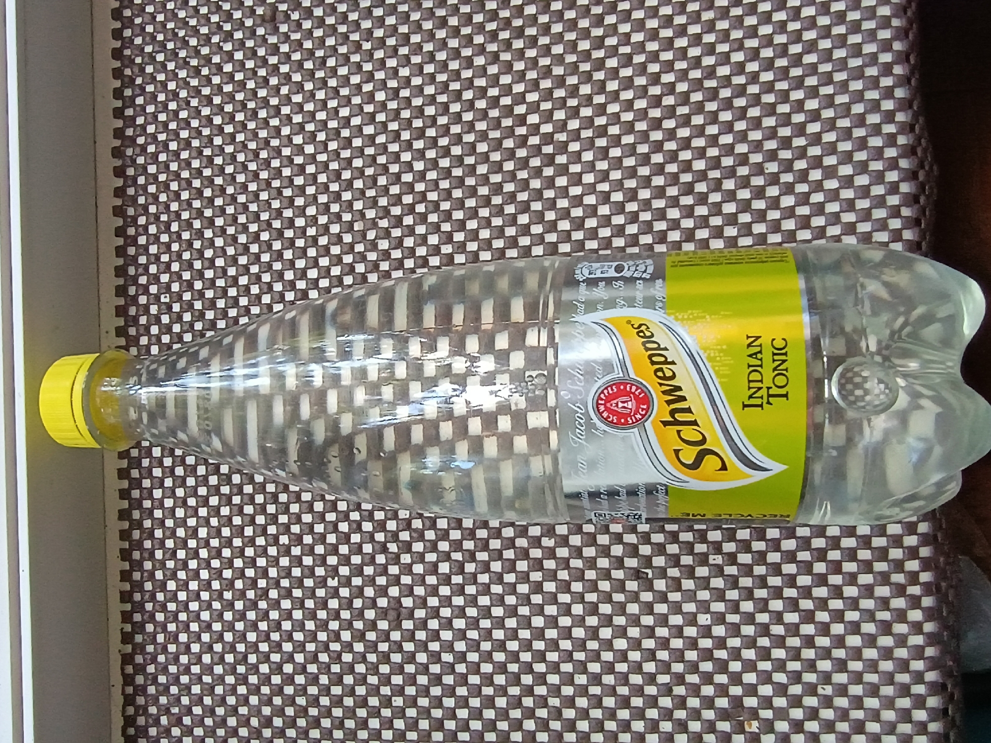 A clear plastic bottle of Schweppes Indian Tonic water lies on a brown and white checkered surface. The bottle is mostly full of clear liquid and has a yellow cap. The label on the bottle is bright yellow and green with white text. Visible text on the label includes "Schweppes," "INDIAN TONIC," and "RECYCLE ME." The bottle appears to be lying horizontally, possibly on a table or mat. The background is a close-up of the checkered surface, and the lighting suggests it could be daytime indoors. There are no people or animals visible in the image. The context of Zaporizhzhia, Ukraine, is provided but not visually evident in the image itself.
