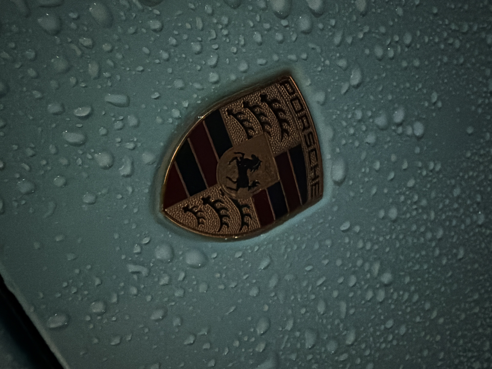 The image presents a forensic close-up of a gold-toned Porsche crest emblem, centered on a light teal-colored vehicle surface. Numerous clear water droplets of varying sizes are distributed across both the emblem and the surrounding paintwork. The shield-shaped emblem features the word "PORSCHE" on a curved banner at its apex. A black rearing horse is prominently displayed within a central, smaller shield. Flanking the central shield are black stylized stag horns on a textured gold background, while the lower sections exhibit alternating red and black vertical stripes. The lighting is subdued, suggesting an outdoor environment, likely a street or parking area in Varna, Bulgaria, during evening or nighttime conditions. No individuals or specific actions are visible within the frame.
