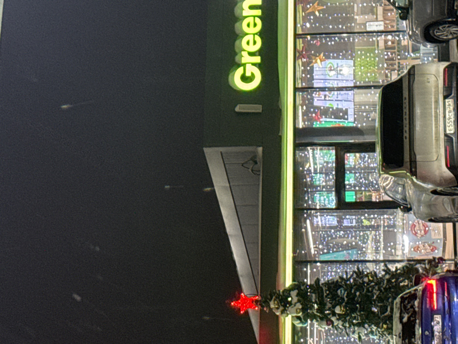 The scene depicts the exterior of a retail establishment at night in Akseno-Butyrki, Russia, during what appears to be a winter or holiday season with light snow visible. The primary subject is a building with a dark green upper facade and a brightly illuminated storefront featuring large glass panels. A prominent neon green sign displaying "I Green" is affixed to the upper facade. An angular, upward-sloping canopy extends from the roofline above the left section of the building.
In the foreground, a snow-dusted Christmas tree, adorned with ornaments and topped by an illuminated red star, stands to the left. Parked vehicles are positioned in front of the store. A blue sedan, partially visible on the left, has its rear lights illuminated and appears to have snow on its trunk. Centrally, a silver or light gray SUV is parked, facing away from the viewer, displaying a Russian license plate "E559CB 761". A portion of a third silver/gray vehicle is visible on the far right. The interior of the store, visible through the glass, is extensively decorated with numerous white string lights and some red star ornaments. No individuals are discernible within the image.
