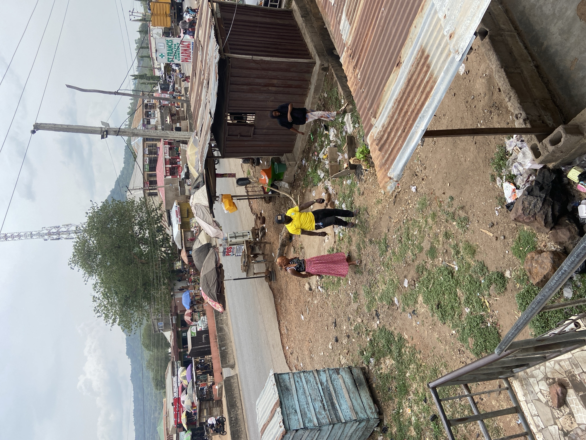 The image depicts an outdoor street scene, likely a bustling marketplace, in an urban or semi-urban setting in Lokoja, Nigeria, captured from an elevated perspective looking down.
The foreground features a weathered blue wooden box-like structure on dirt ground, next to a metal railing. The ground throughout the scene is primarily unpaved dirt with sparse green vegetation and significant amounts of scattered litter, including plastic bags and general debris.
Several people are visible. In the midground, a person wearing a bright yellow t-shirt, dark pants, and a dark cap stands on the dirt. Nearby, a woman in a long, flowing pink or red skirt and a dark top is also present. Closer to a dark brown corrugated metal structure, another person in a black top and light-colored patterned pants is seen. Further down the street, numerous other individuals are visible, many congregating near market stalls or under colorful umbrellas providing shade.
The street itself, partially paved, runs horizontally across the midground, lined with various commercial structures. These include makeshift stalls and more permanent buildings, many featuring corrugated iron roofs. Visible signs include "ST. FRANCIS HEALTH CONSULT... PHARMACY" on a white building, and "POS SERVICE" on a smaller sign. Another partially visible sign reads "JUNIOR HOME ESSEN...". Utility poles with power lines crisscross the upper part of the scene. A large green-leafed tree stands prominently on the left, and a cellular communication tower is visible in the far distance. Rolling hills or mountains form the backdrop under an overcast sky.
The scene portrays daily life and commerce, with people moving or standing, suggesting a regular market day. The time appears to be daytime, characterized by a soft, diffused light consistent with cloudy weather.