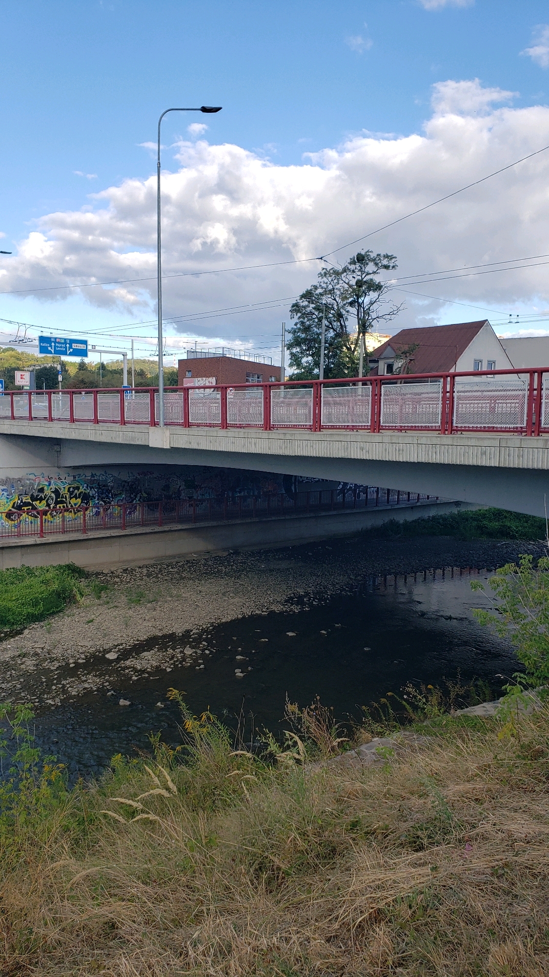 The image shows a view of the Sekčov river and a pedestrian bridge in Prešov, Slovakia. The bridge has a red railing and is covered in graffiti underneath. The river is shallow with a rocky bed. On the bank of the river, there is dry grass and some green vegetation. In the background, there are buildings, trees, a street light, and traffic signs. The sky is blue with scattered clouds.