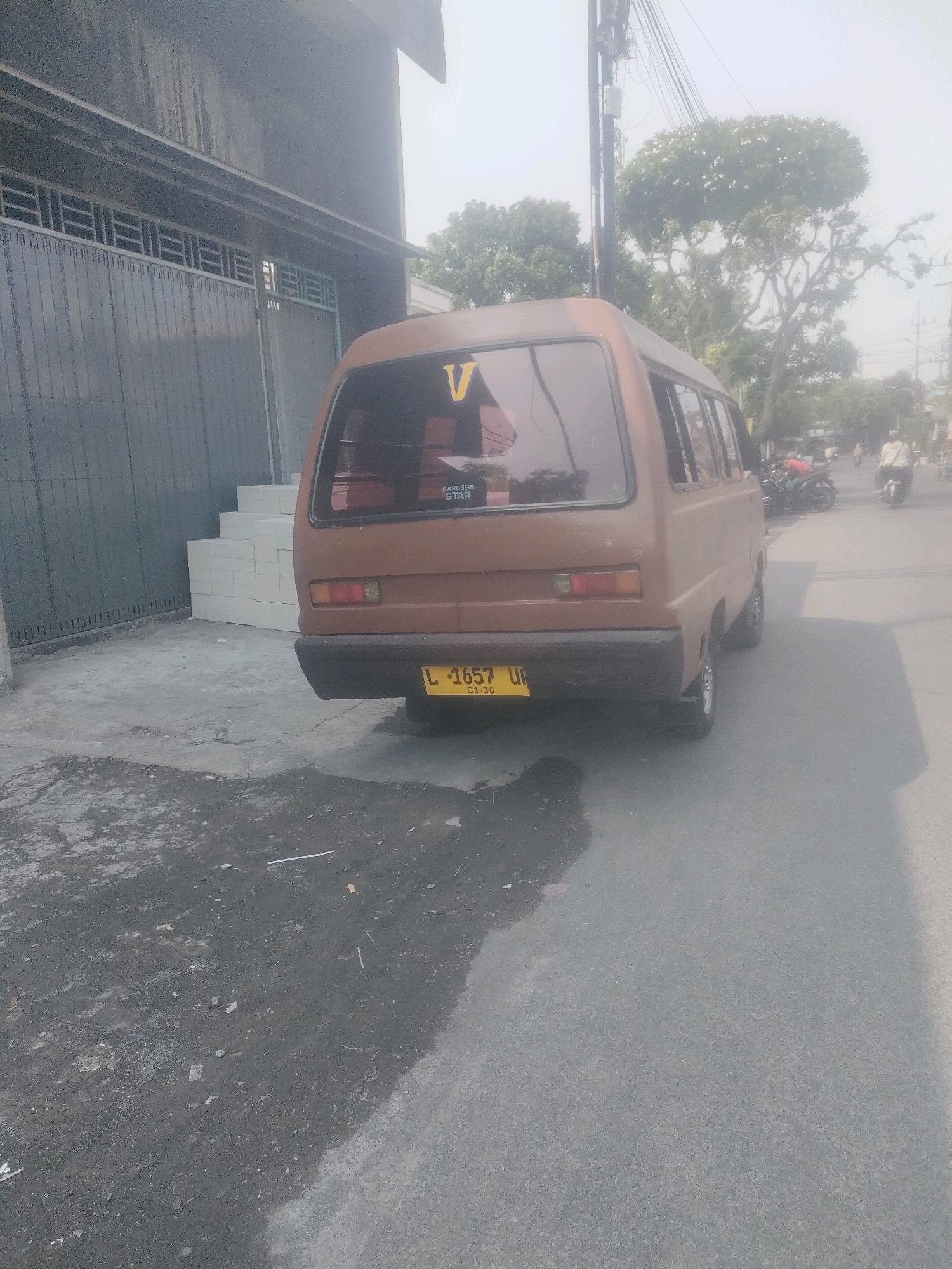 In Surabaya, Indonesia, a brown van is parked on an asphalt road next to a grey building. The building has a metal gate and a stack of white bricks on its side. The van has a yellow license plate that reads "L 1657 UF" and the number "01-20" underneath. There are trees and power lines in the background, and several motorcycles and a person on a motorcycle are visible further down the road. The sky is hazy and light.