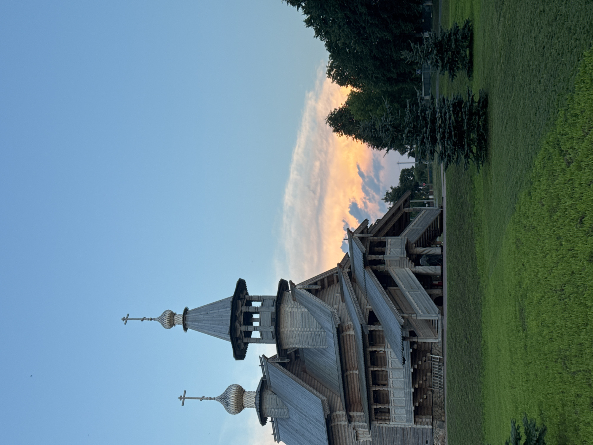 An outdoor, medium shot captures a wooden, traditional architectural style structure with multiple levels and tiered roofing, topped with onion domes and crosses, set against a twilight sky with wispy clouds tinged with orange and pink. The building's exterior is constructed from weathered wooden logs and features intricate wooden carvings and latticework on its balconies and staircases. Two pine trees stand in the foreground on a manicured green lawn, with a line of darker trees forming the background to the right. The sky is a gradient of light blue to a softer hue near the horizon, indicating early evening. No people are visible in the frame. The overall scene suggests a historical or cultural site, possibly a church or museum, in the Нёманскі сельскі Савет, Belarus.