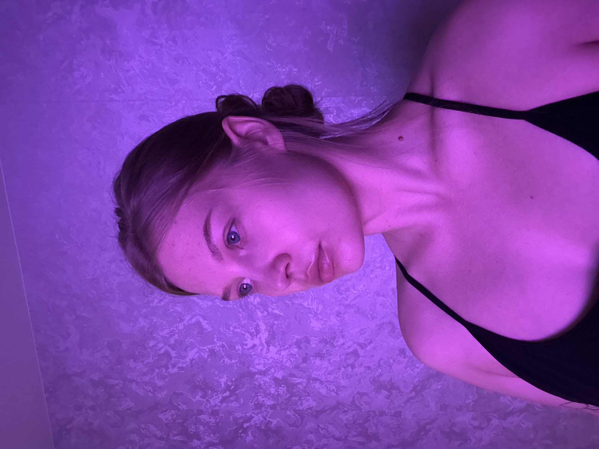 A young woman is depicted from the head to the upper torso, set against a textured, light-colored wall. The entire scene is bathed in a dominant, pervasive purple light, suggesting an artificial lighting setup or a specific mood. The image is rotated 90 degrees counter-clockwise, presenting the woman's head on the left side of the frame and her body extending downwards.
She has light hair, pulled back into a bun. Her blue eyes are open and directed towards the viewer, or slightly past the camera. Her facial expression appears neutral to slightly pensive, with her lips gently parted. She is wearing a dark, likely black, spaghetti-strap top or garment. Notable physical details include a mole on her neck/shoulder area.
The background wall has a distinct, mottled texture, which is subtly highlighted by the purple illumination. No specific activity is taking place beyond posing for the photograph. The intense, monochromatic lighting makes it difficult to determine the time of day, but it implies an indoor setting, possibly at night. There are no visible location cues within the image itself to suggest the provided context of Chaikovsky, Russia. No text is visible in the media file.