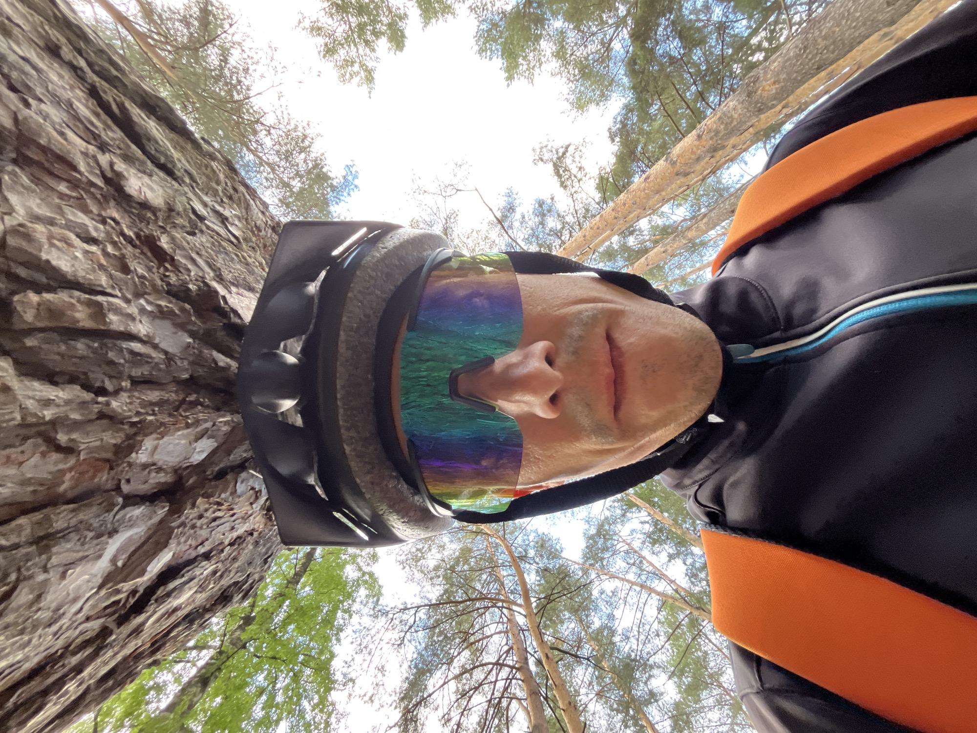 A low-angle, close-up photograph captures a person in a forest setting in Chaikovsky, Russia. The individual, seen from the nose downwards, wears a dark cycling helmet and large, rainbow-reflective sunglasses that obscure their eyes. They are dressed in a black long-sleeved athletic top with subtle blue and white detailing on the zipper, and a prominent bright orange strap, likely from a backpack or safety harness, crosses their shoulder.
The camera's perspective is directed sharply upwards, showing a dynamic view of the surroundings. On the left side of the frame, a large tree trunk with deeply textured, rugged bark fills a significant portion of the image, suggesting an old or mature tree. The background is dominated by the canopy of a pine forest, with numerous tall, slender tree trunks and green foliage silhouetted against a bright, clear sky. The abundant natural light indicates it is daytime, likely during clear weather.
The person's attire and the natural environment suggest engagement in an outdoor activity such as cycling, mountain biking, or hiking. No visible text is present in the image.