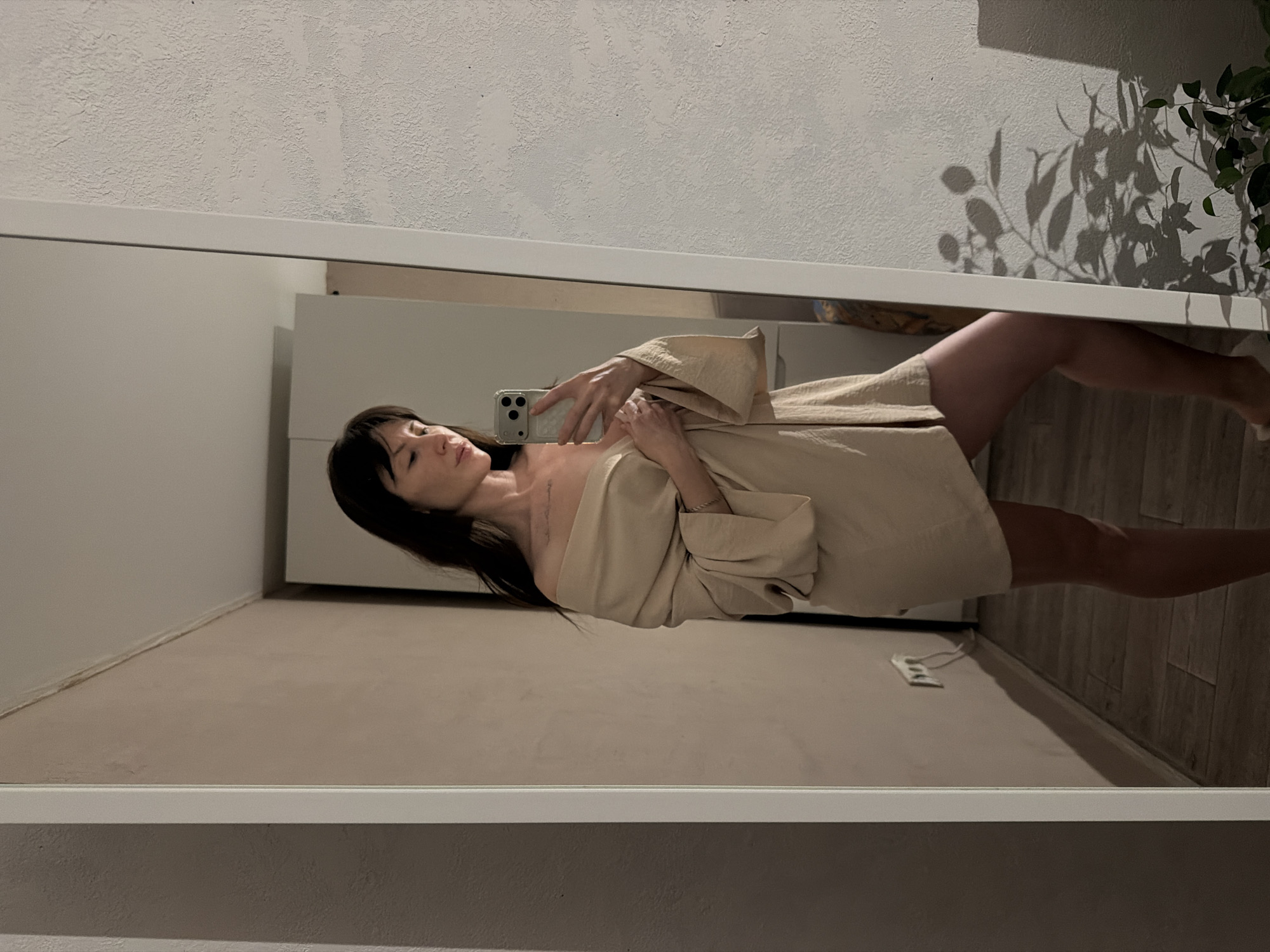 A woman is reflected in a mirror. She is holding a cell phone, taking a picture of herself. She is wearing a tan robe. The location is likely indoors, as there is a wall and a plant visible in the reflection. Based on visual elements, the location in the city of Perm, Russia is unknown.